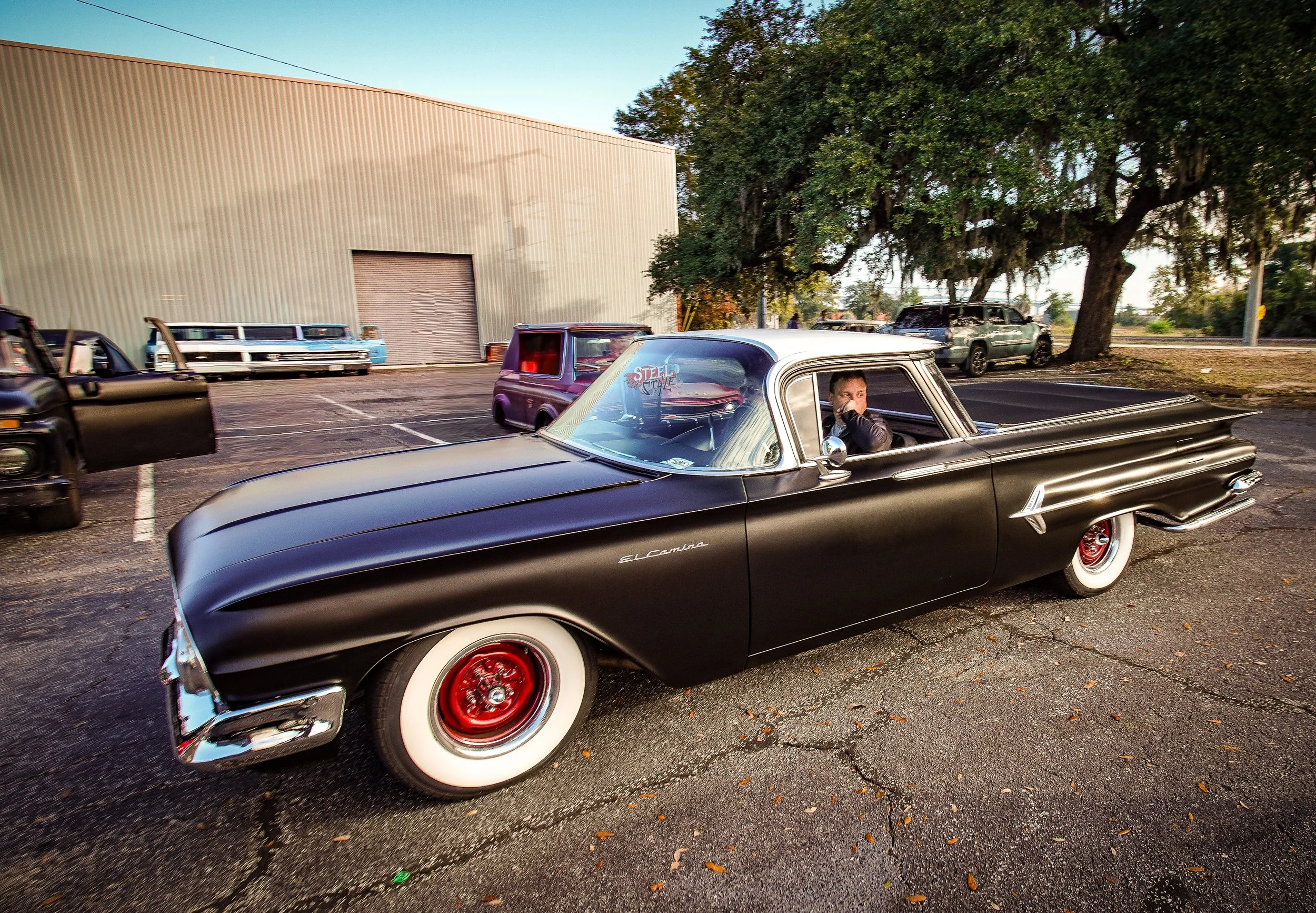 A vintage black and white two-tone 1950s Chevrolet car parked in an outdoor lot near several other classic cars, with a large tree and industrial building in the background.