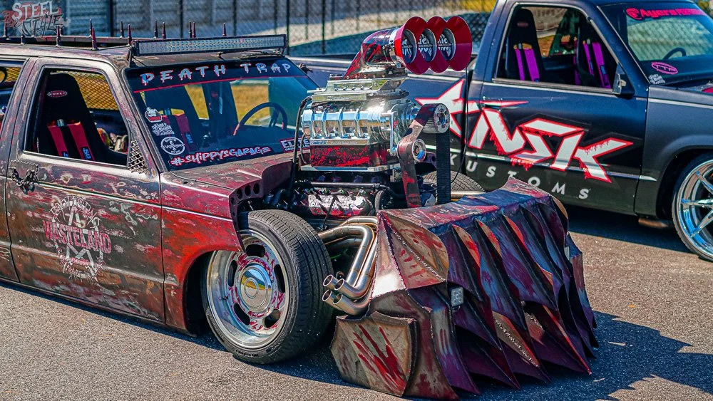 A heavily modified racing pickup truck with a large supercharger sticking out of the hood, painted in a red and black, distressed style, and featuring a massive front plow attachment. The truck has various decals, including 'Death Trap' on the windsh