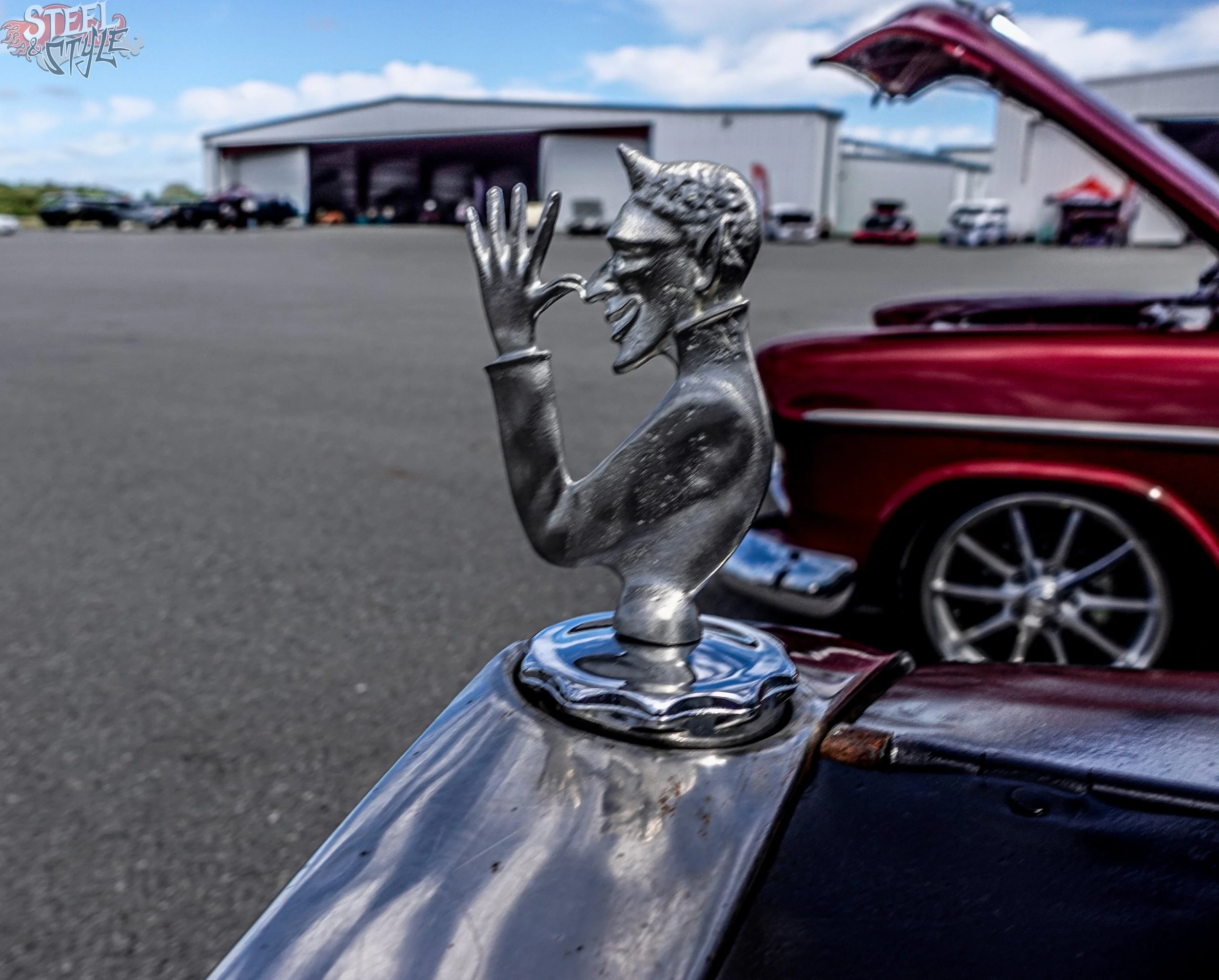 Close-up of a shiny chrome hood ornament representing a cartoonish character with a big smile, raised hand, glasses, and spiky hair on the front of a red vintage car, with a parking lot and industrial building in the background.