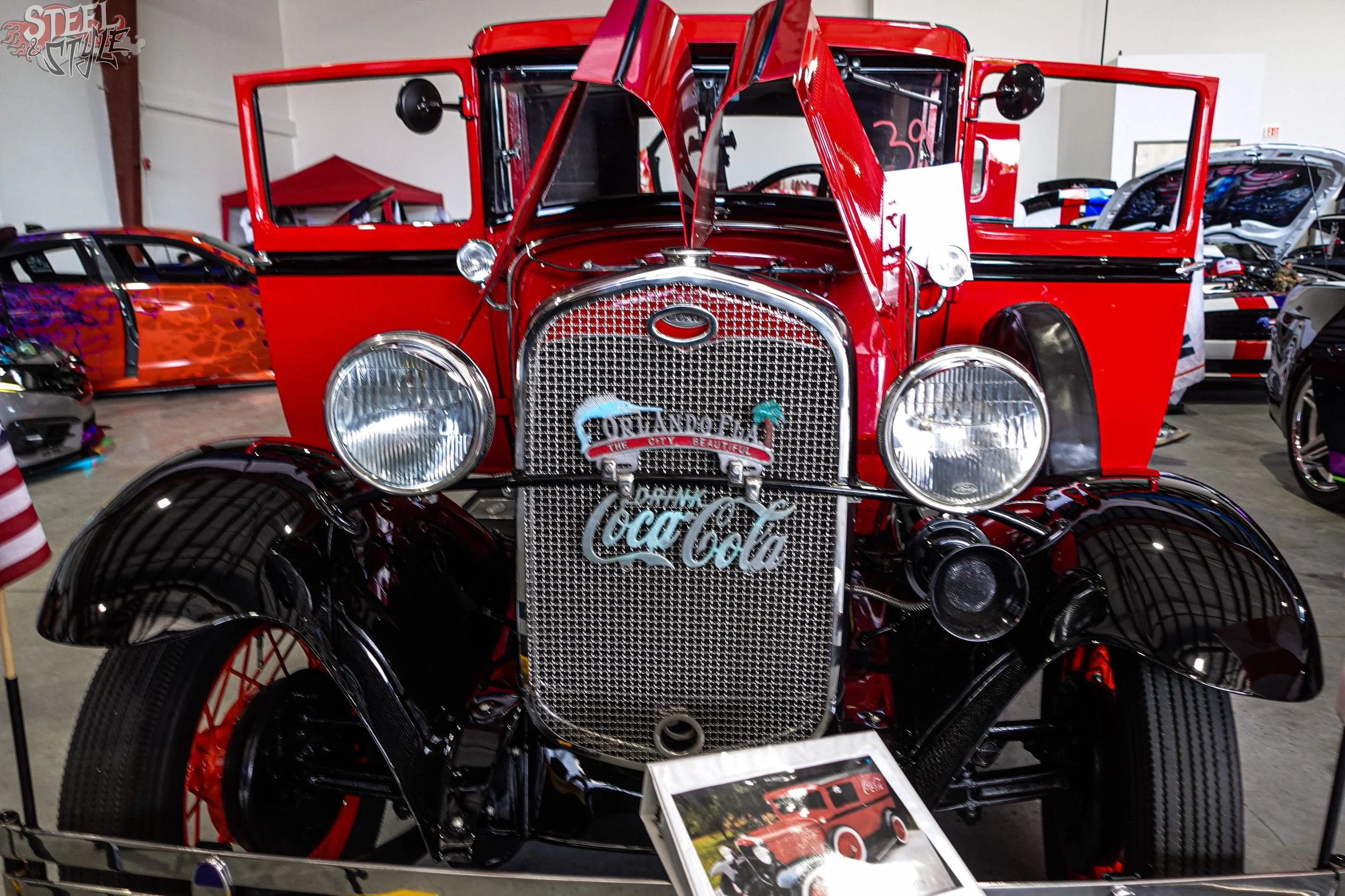 A vintage red Ford Model T car with the hood open, displayed at an indoor exhibition. The front grille has a Coca-Cola sign attached, and the car is surrounded by other vehicles and exhibit information.