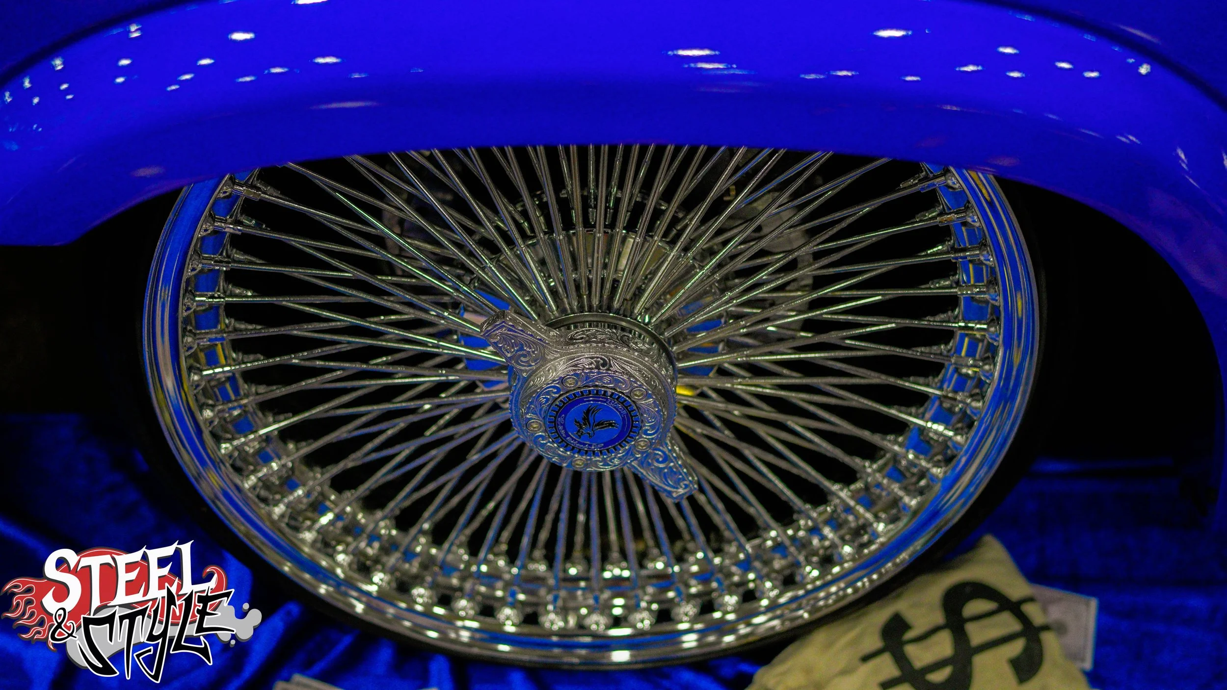Close-up of a shiny wire-spoke wheel of a vintage car, with a decorative center cap featuring a phoenix emblem, under blue lighting.