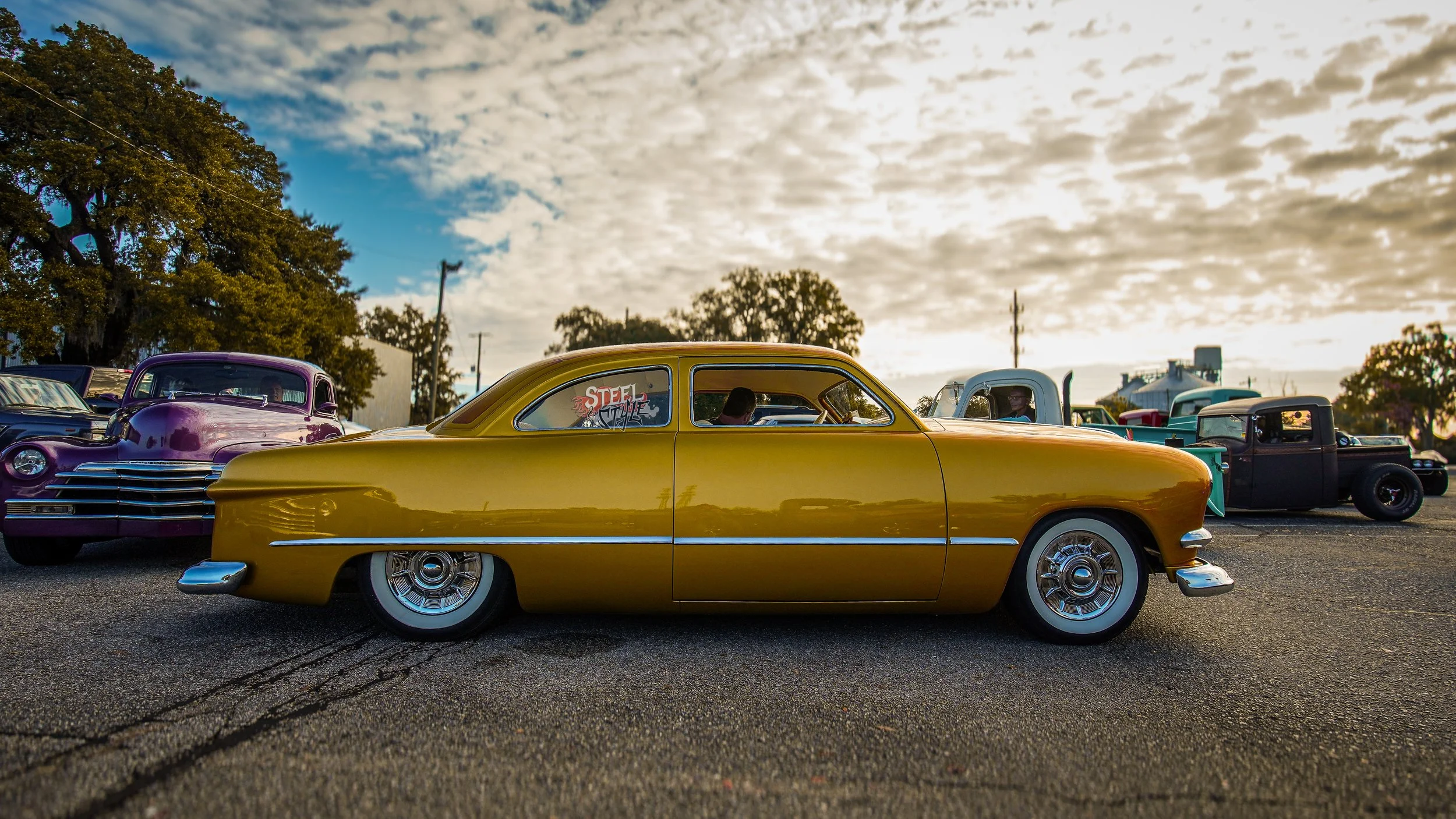 A yellow vintage car parked among other classic cars outdoors during sunset, with cloudy sky and trees in the background.