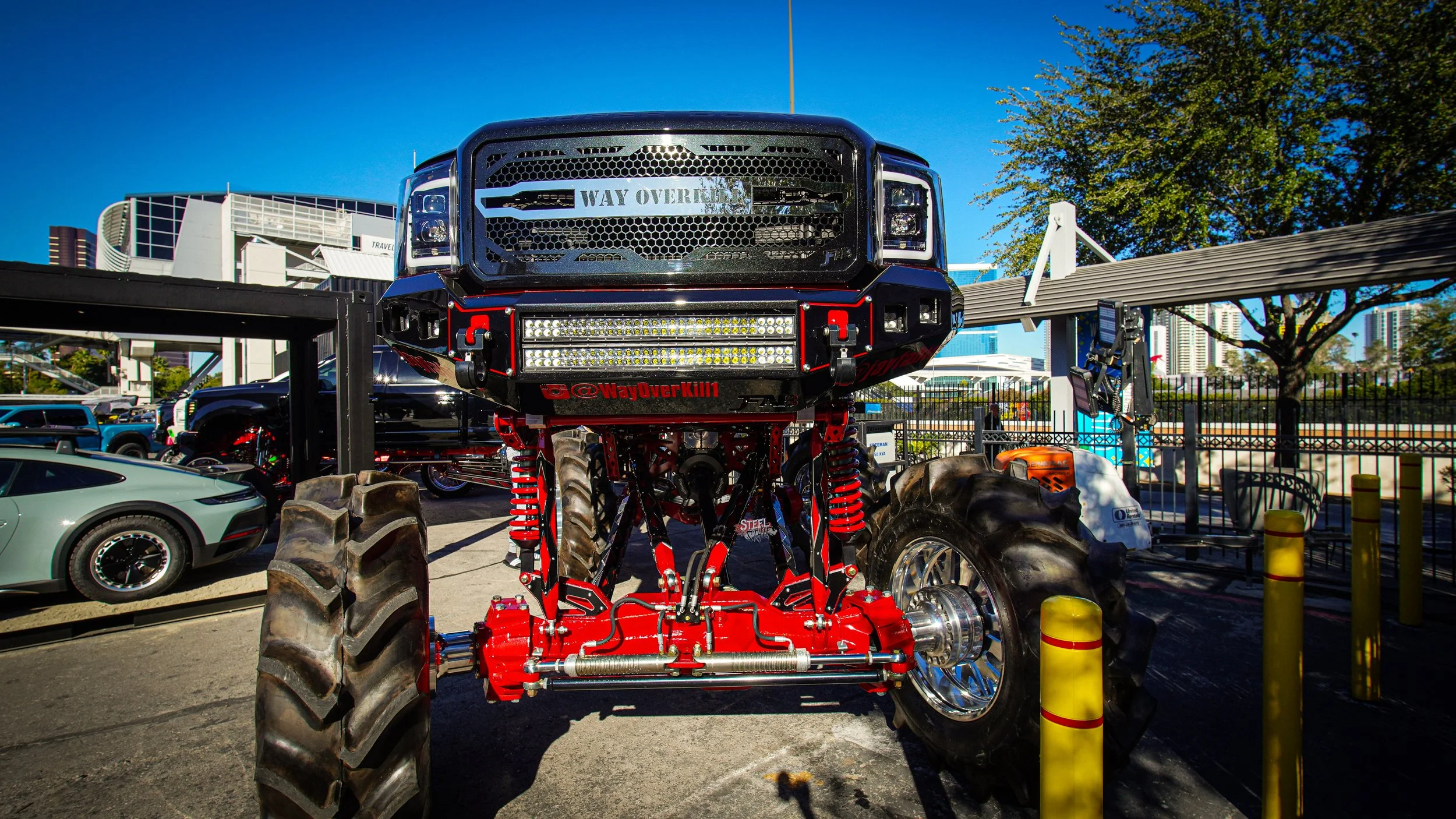 A large monster truck with red and black coloring, big tires, and aftermarket parts, parked in an outdoor lot with other vehicles and a city skyline in the background.