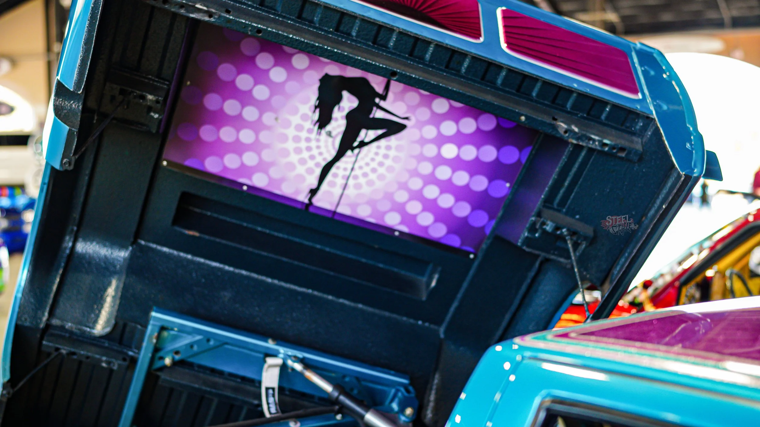 A carnival game with a digital screen displaying a silhouette of a woman pole dancing against a purple polka-dot background, mounted on a blue structure at an amusement park or fair.