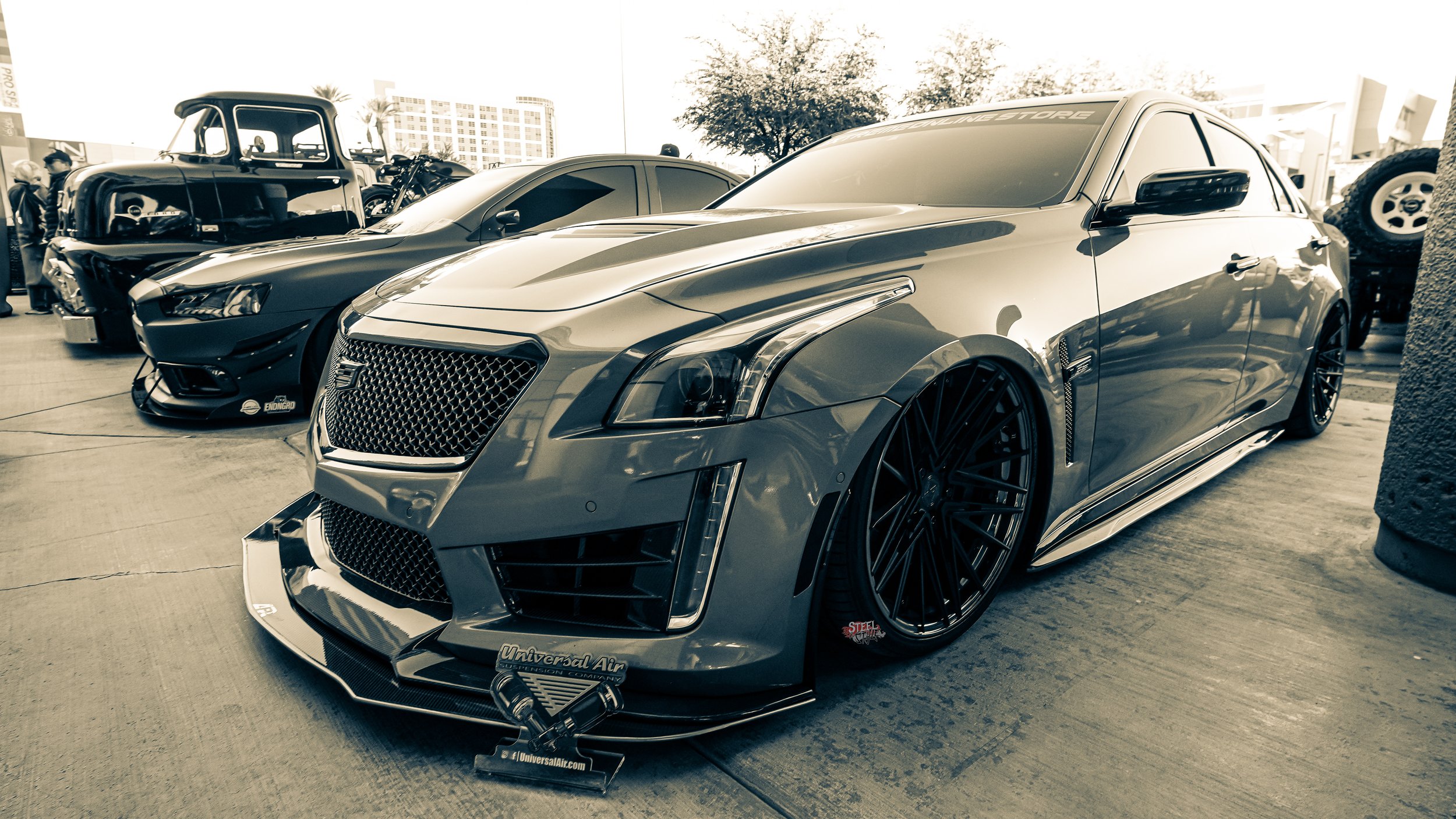 A line-up of customized cars at a car show, featuring a sleek, low to the ground grey sports car with large black rims in the foreground, and other classic and muscle cars in the background.