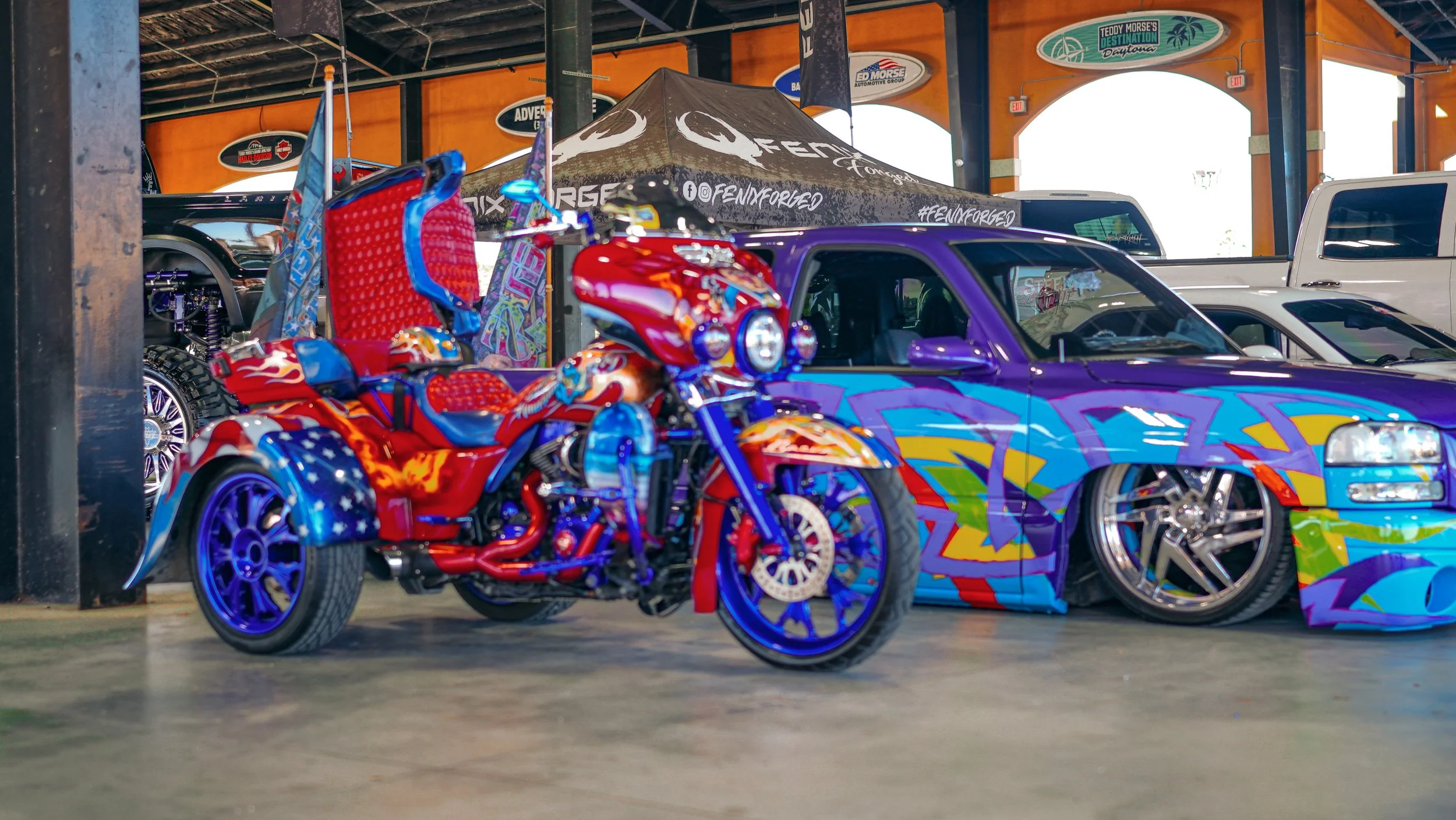 A colorful motorcycle with patriotic red, white, and blue flames, and a trike seat, parked next to a graffiti-style purple and blue car inside a venue with orange walls and signs.