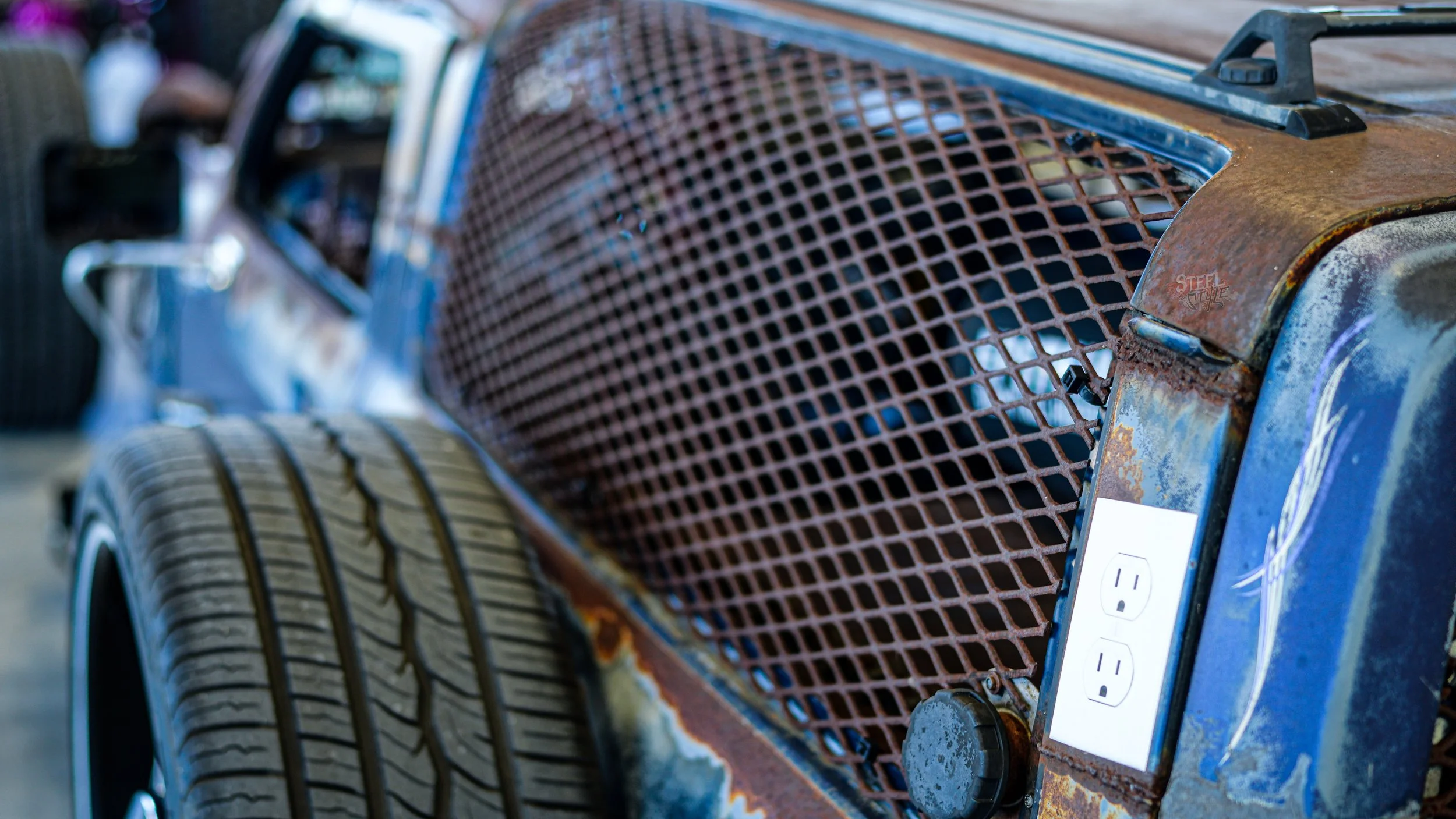 Close-up of an old, rusty vintage car with a metal grille and a plug socket on the front fender, showing signs of wear and rust.