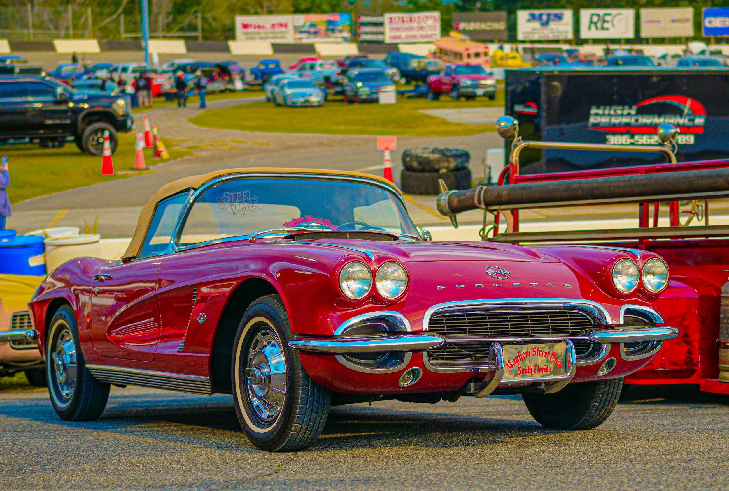 A red vintage Chevrolet Corvette with a beige soft top parked at a car show, surrounded by other classic cars and vehicles in the background.
