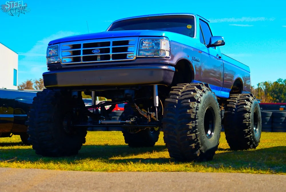 A blue Ford F-150 truck with oversized off-road tires and suspension is shown on grass, lifted high above the ground.