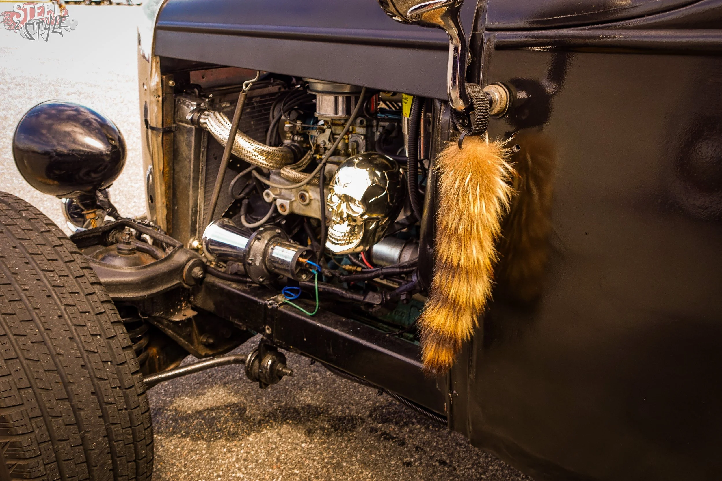 Close-up of a vintage car's engine bay, featuring a skull decal, shiny metallic parts, black hoses, and an orange furry material on the side.
