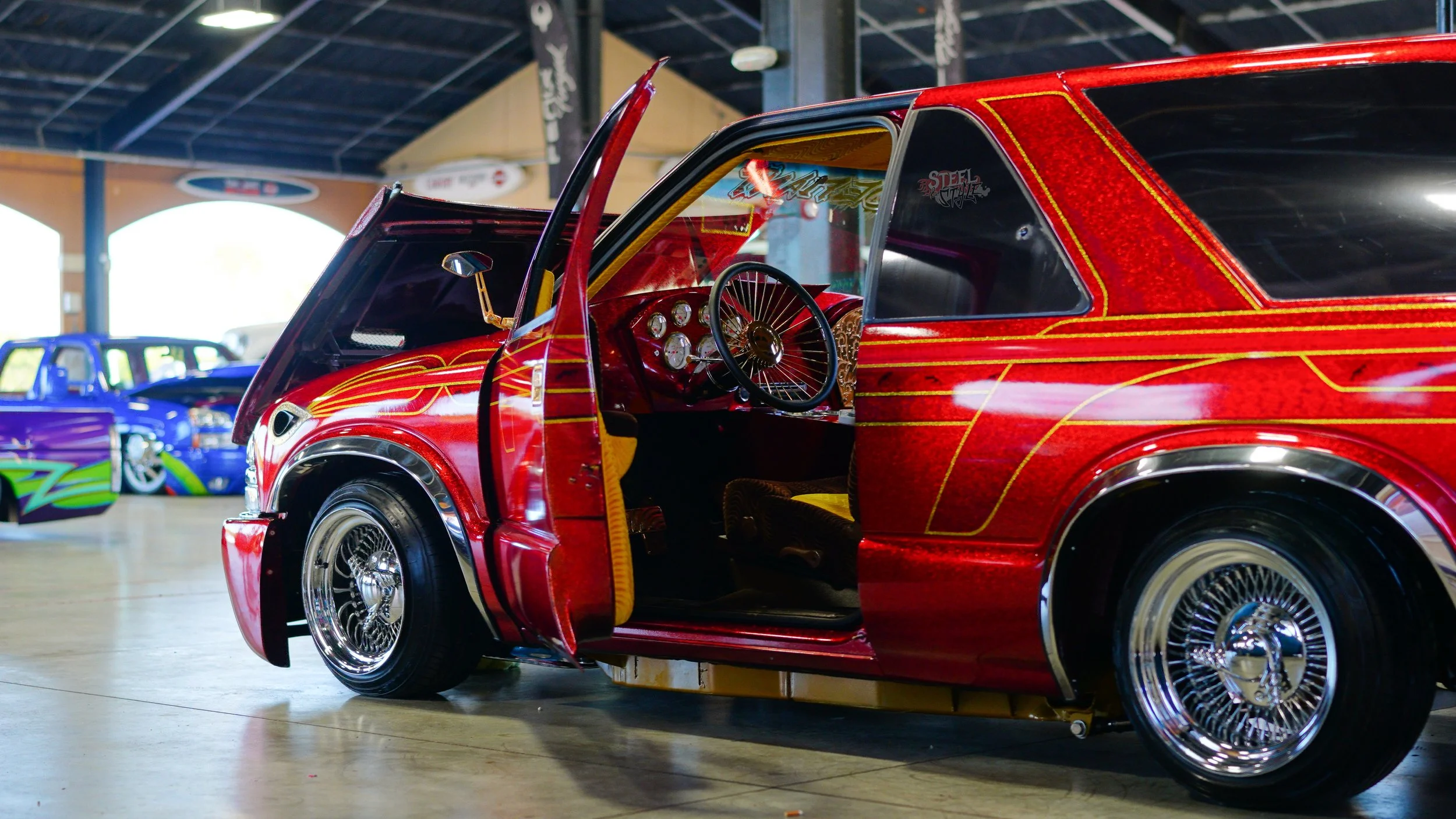 A red custom-designed race car with gold accents and chrome wire wheels, parked indoors at a car show, with another purple and green race car visible in the background.