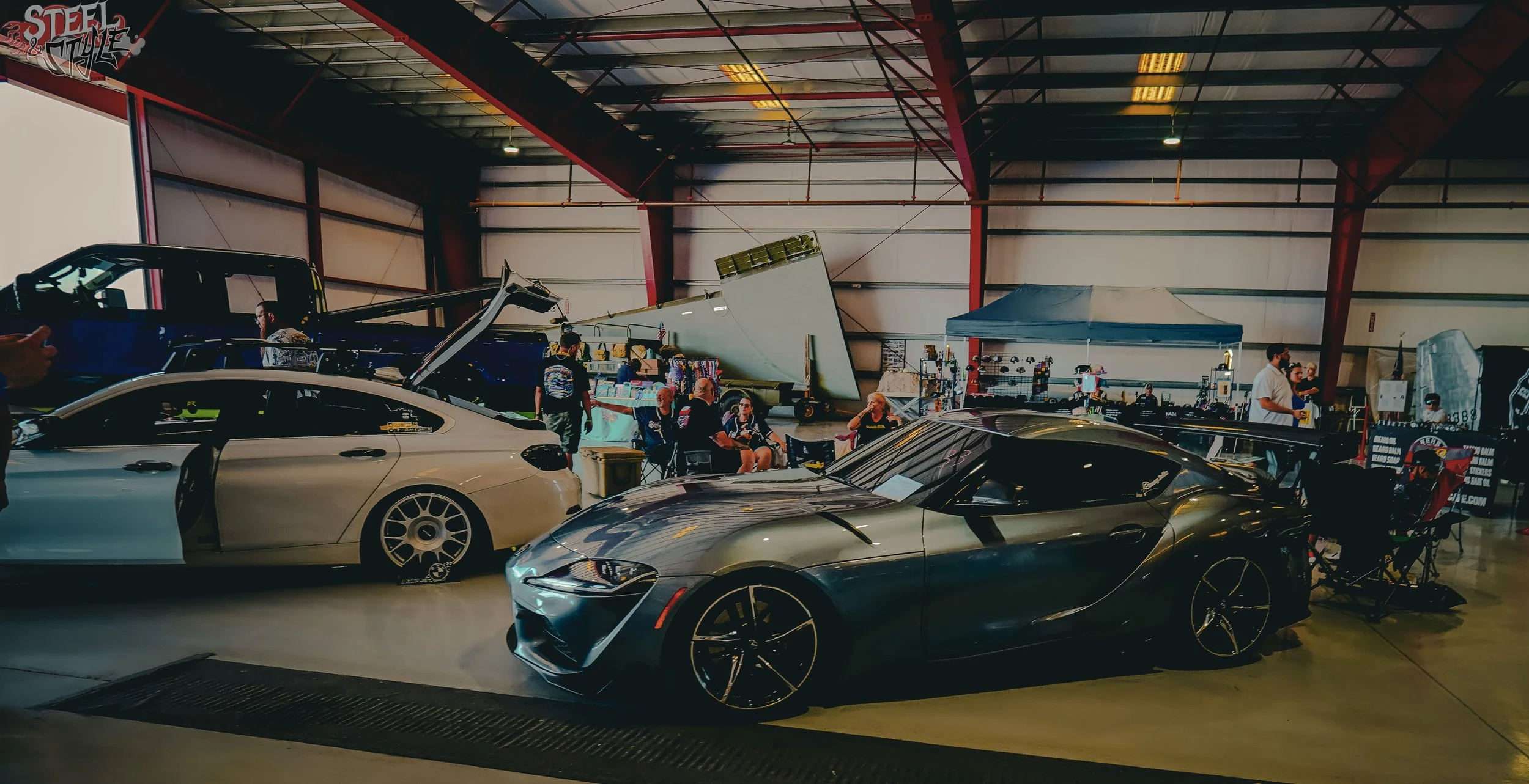 An indoor car show with various luxury and sports cars, vendors, and attendees, under a large metal-roofed structure.