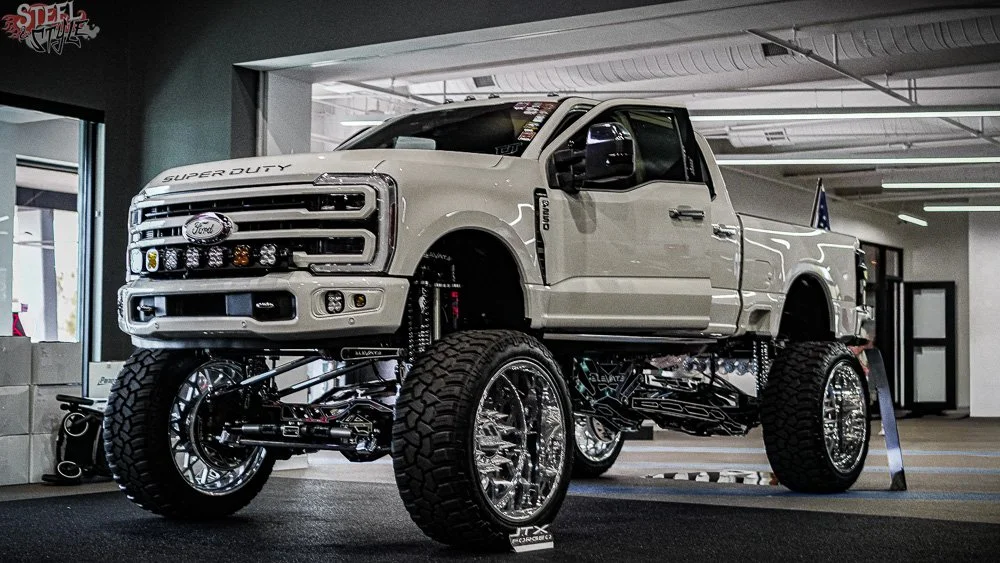 A highly customized Ford Super Duty pickup truck with oversized tires, lift kit, and chrome rims on display inside a dealership or showroom.