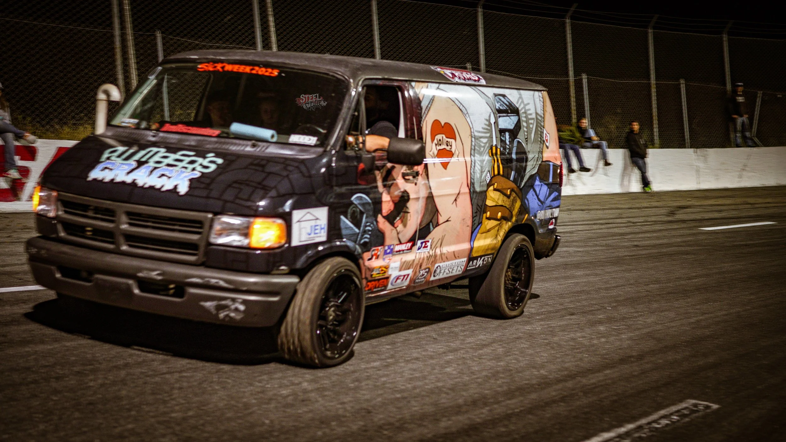A small racing van with colorful cartoon-style artwork on the side, driving on a racetrack at night.