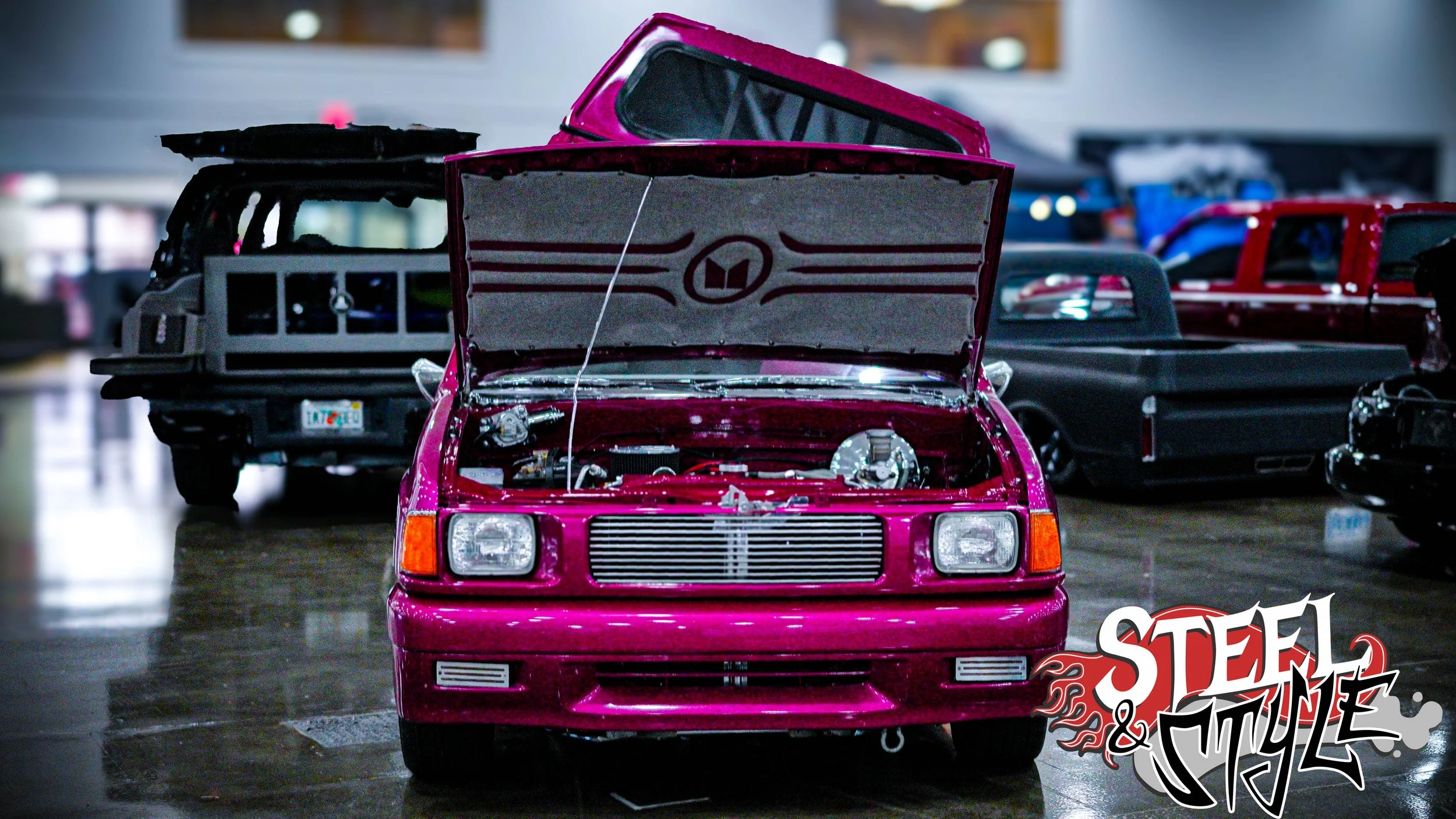 A pink classic car with its hood open, displaying the engine at an indoor car show, with other vintage vehicles in the background and the Steel & Style logo in the bottom right corner.