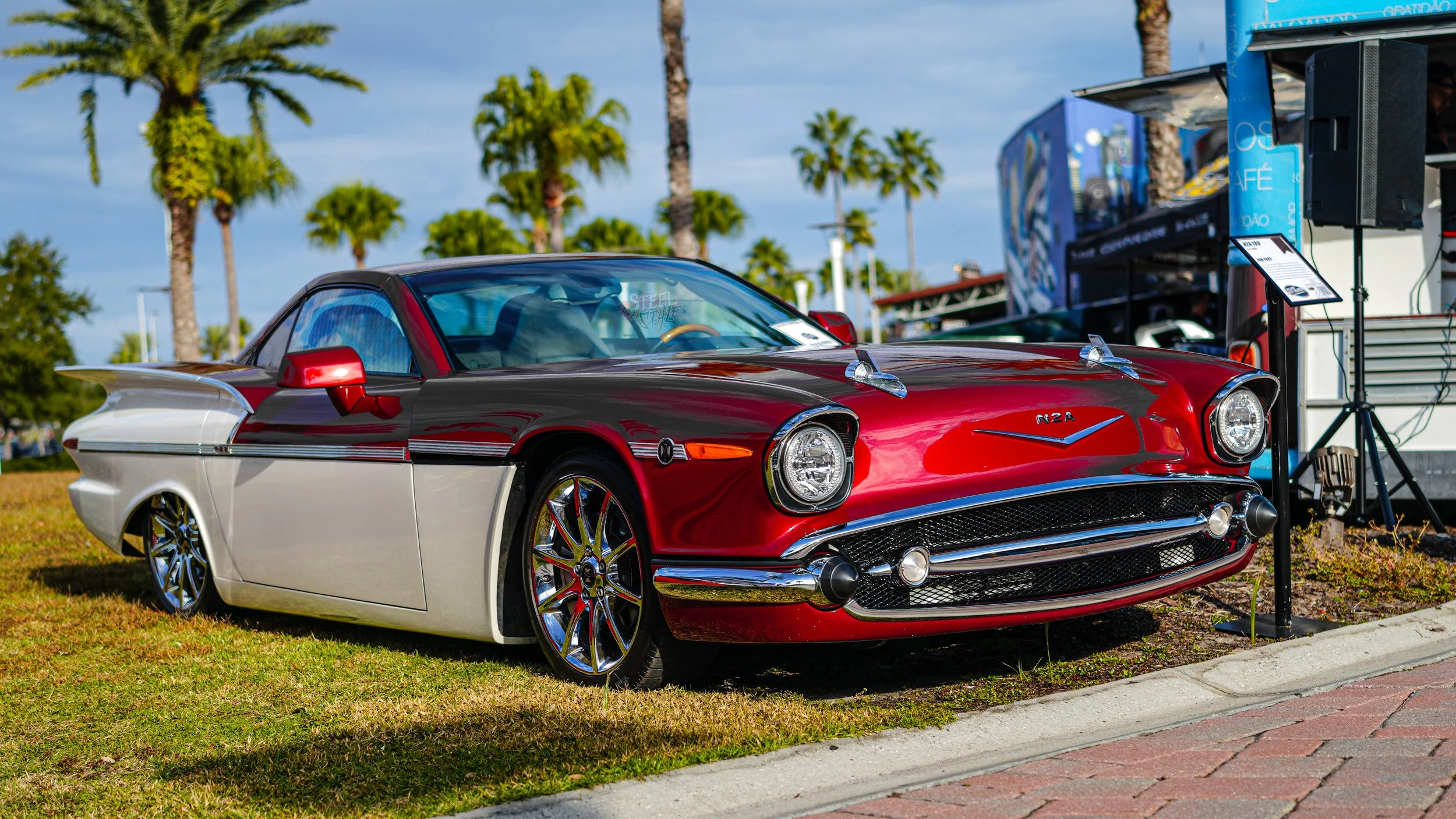 A vintage red and white Chevrolet Corvette with chrome accents parked on a grassy area at an outdoor car show with palm trees in the background.