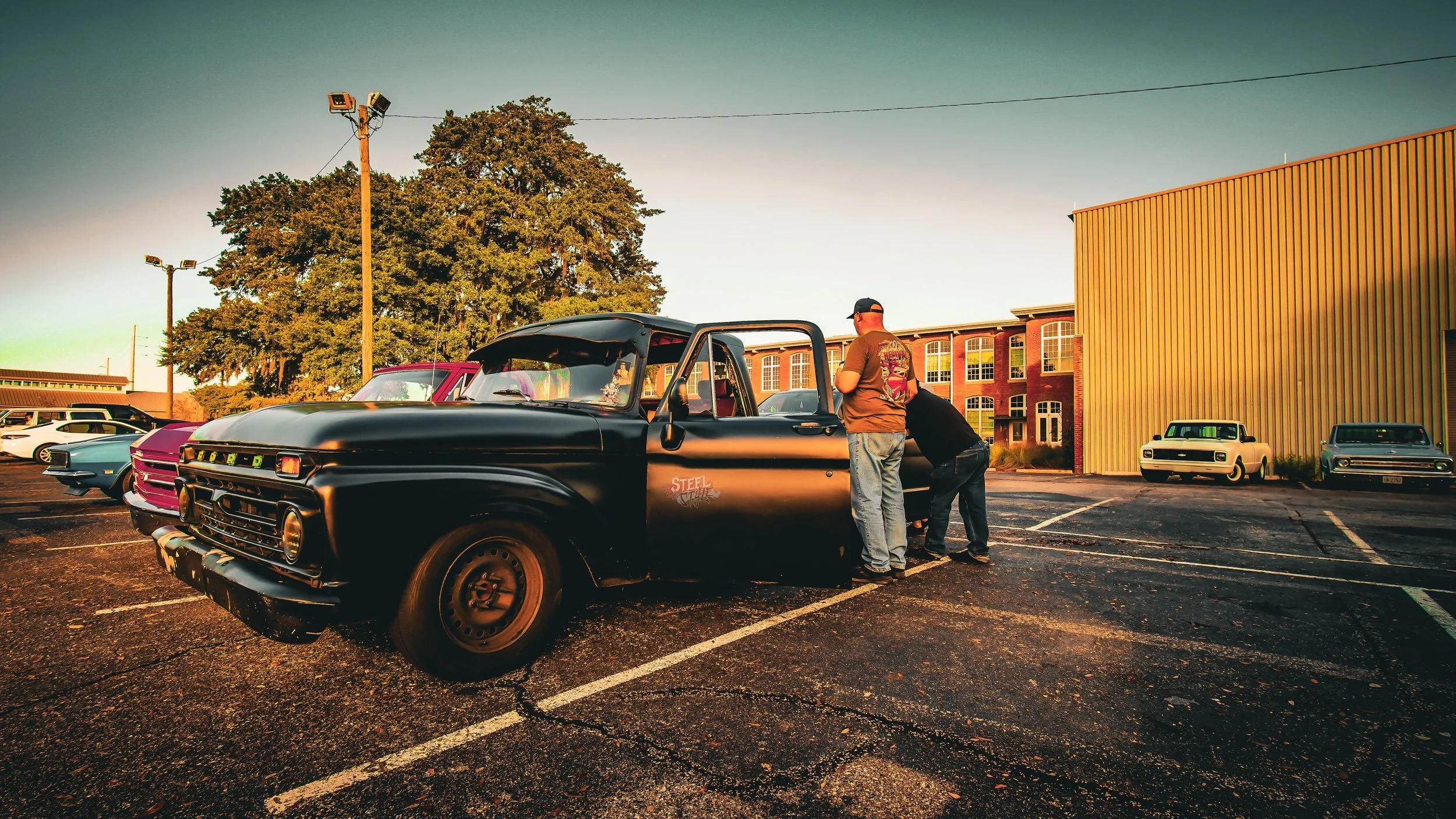 A vintage black pickup truck parked in a lot with two men inspecting its engine. Other classic cars are visible in the background, with a large tree and a building with brick and yellow siding.