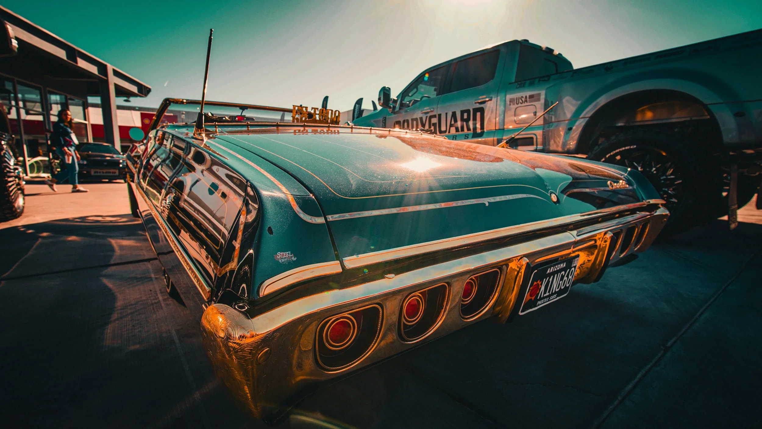 A vintage blue car with a shiny chrome bumper and taillights, parked outdoors with other vehicles and people nearby, under a clear sky during sunset.