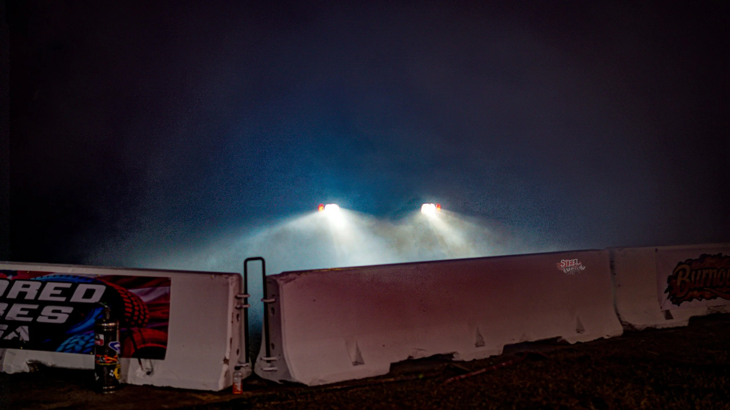Nighttime dirt track with bright headlights illuminating the dust, with barriers and posters on the side.