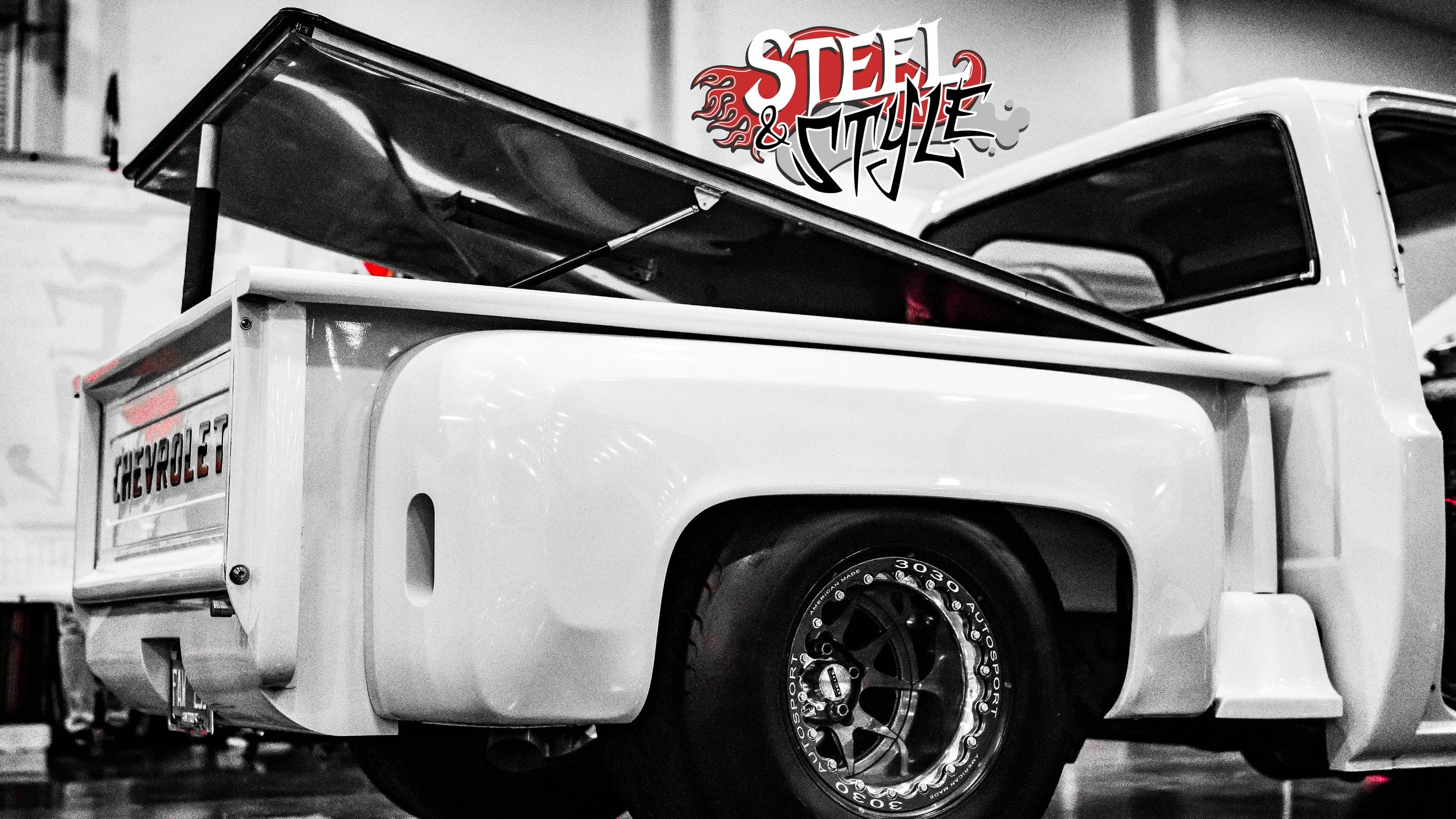 Black and white photograph of a vintage white pickup truck with Chevrolet branding on the tailgate. The truck has racing wheels and a raised bed lid. "Steel & Style" graffiti-style logo is visible in the background.