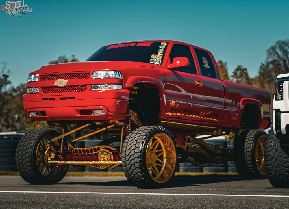 A lifted red Chevrolet pickup truck with large off-road tires and gold accents parked outdoors during daytime.