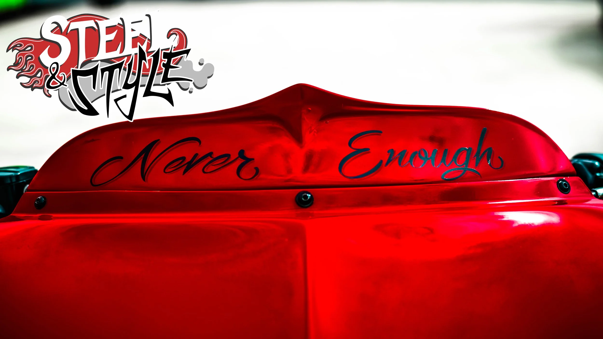 Close-up of a red motorcycle with the words "Never Enough" written on it in cursive. A logo in the top left corner reads "Steel & Style" with a flame design.