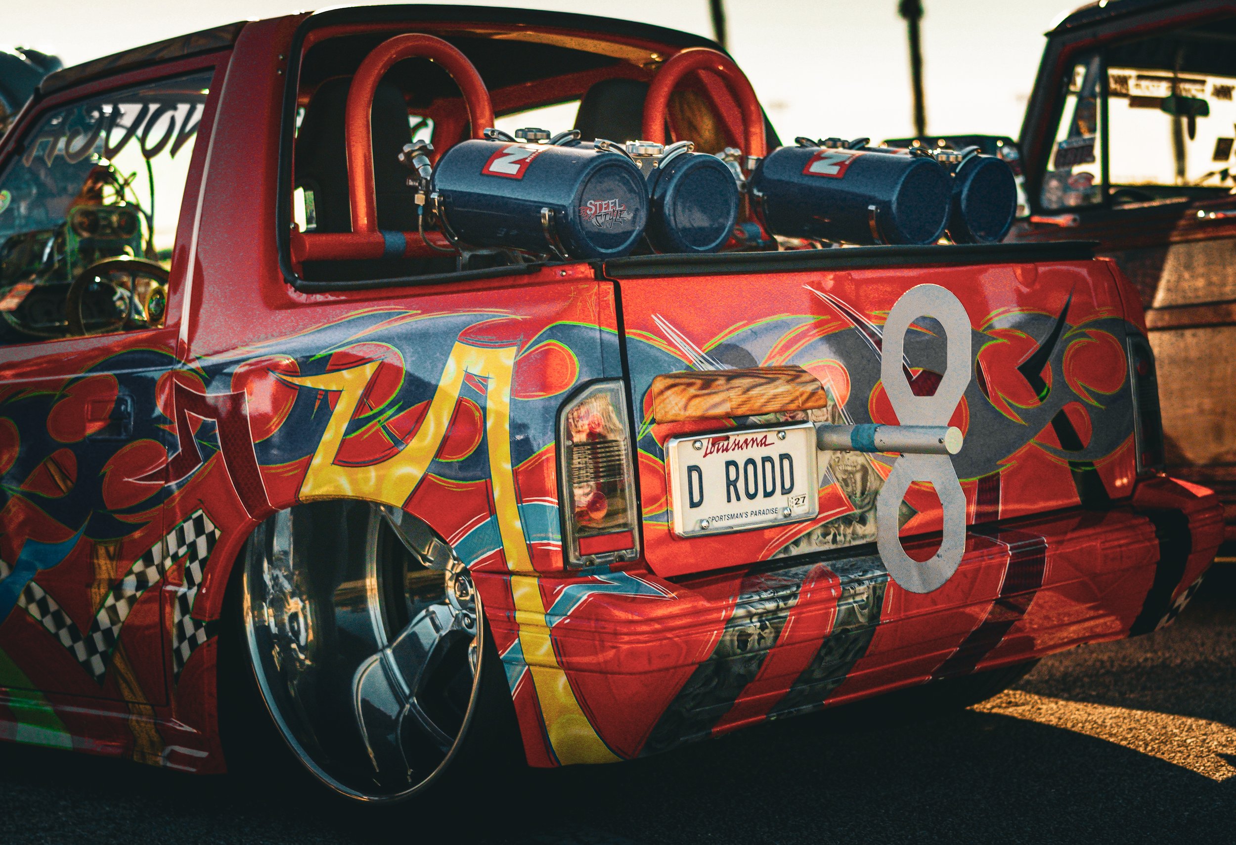 A colorful race car with vibrant graphics, featuring a Louisiana license plate reading D RODD, low-profile tires, and a custom paint job with flames and checkered patterns.