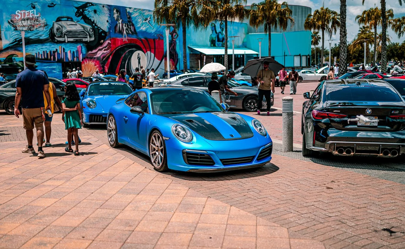 Blue Porsche 911 parked among performance cars at the Velocity Rally outside Dezerland Park in Orlando, with spectators, palm trees, and a colorful automotive mural in the background.