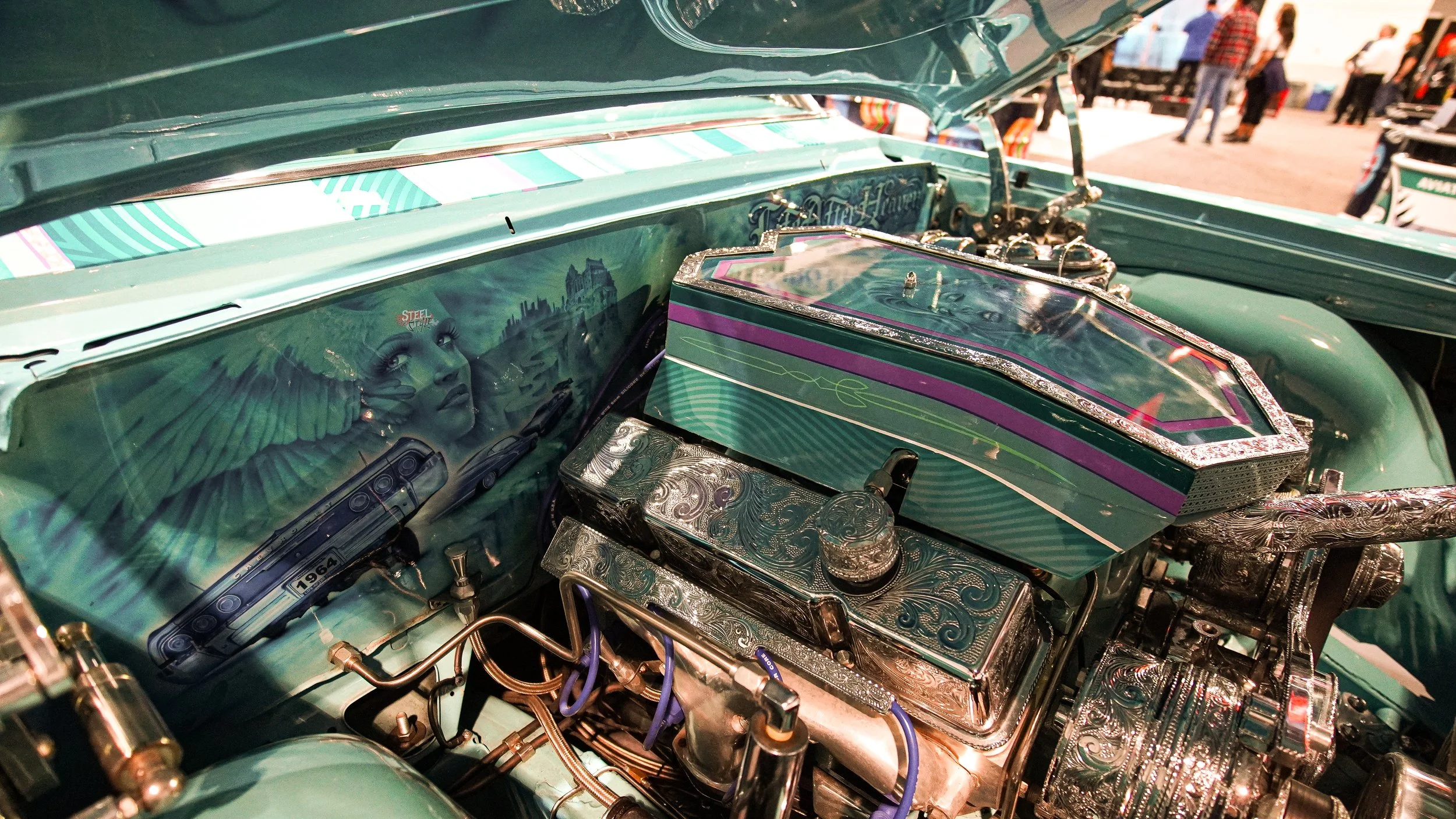 Close-up of a vintage car engine with ornate engravings, painted in turquoise with purple accents, at an automotive show.