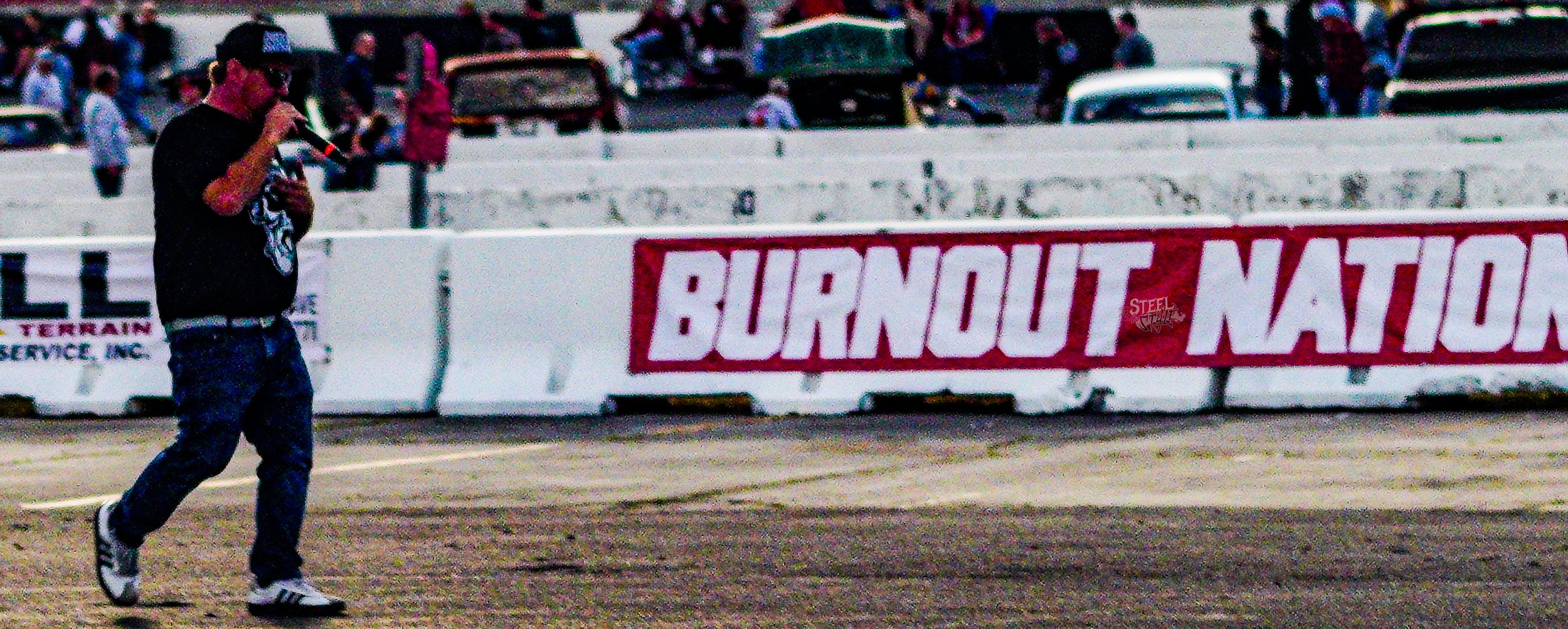 A man in a black t-shirt, jeans, and sneakers walking on a dirt racetrack while speaking into a microphone. Behind him is a white barrier with a red and white banner that reads "BURNOUT NATION," and there are cars and spectators in the background.