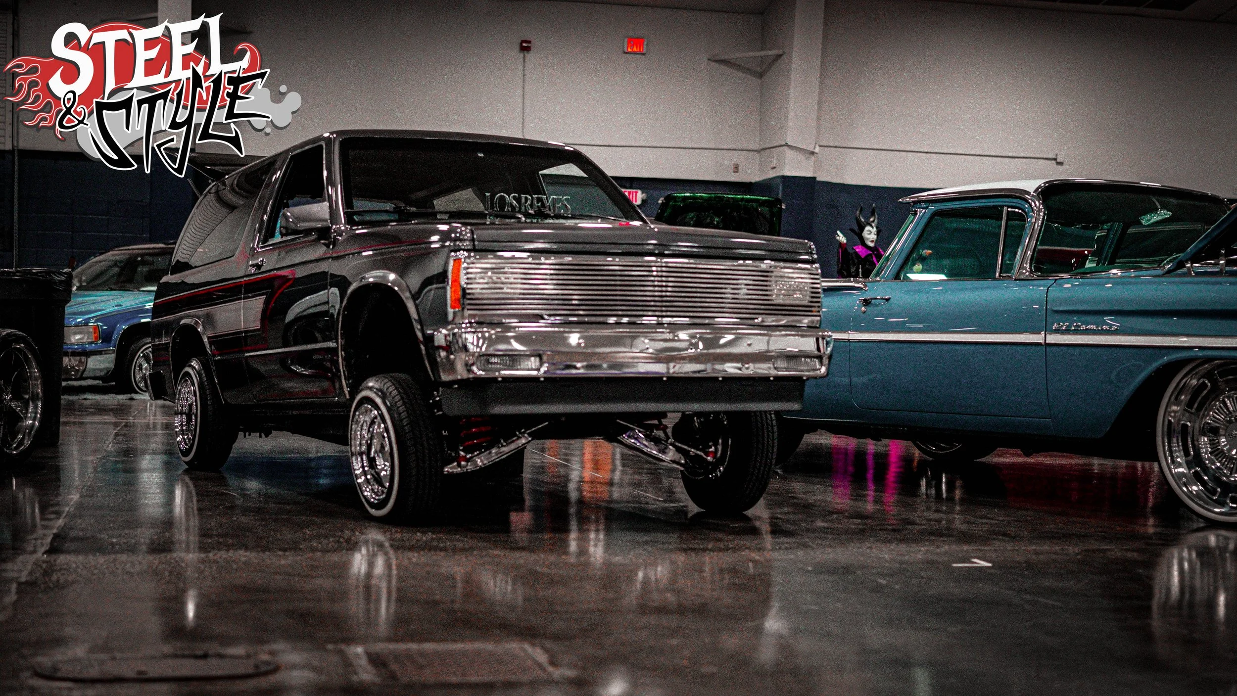 Multiple classic and custom cars, including a black and chrome lifted truck and a blue vintage car, displayed indoors at a car show, with a sign reading 'Steel & Style' in the upper left corner.