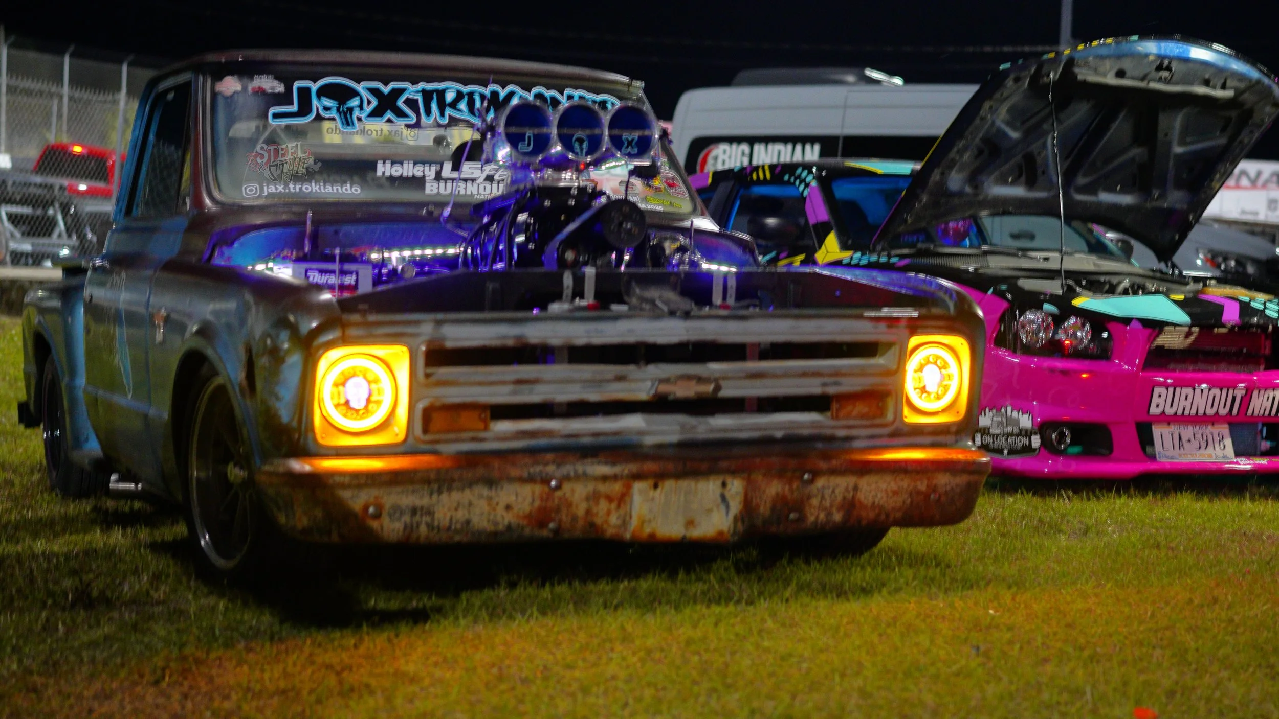 A rusty old pickup truck with bright yellow headlights and a heavily modified engine with purple accents, parked on grass at night, with another customized car in the background.