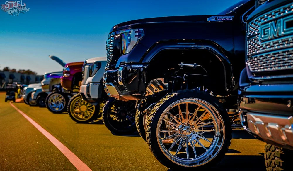 Line of large, customized trucks parked on a grassy field with a clear blue sky in the background.