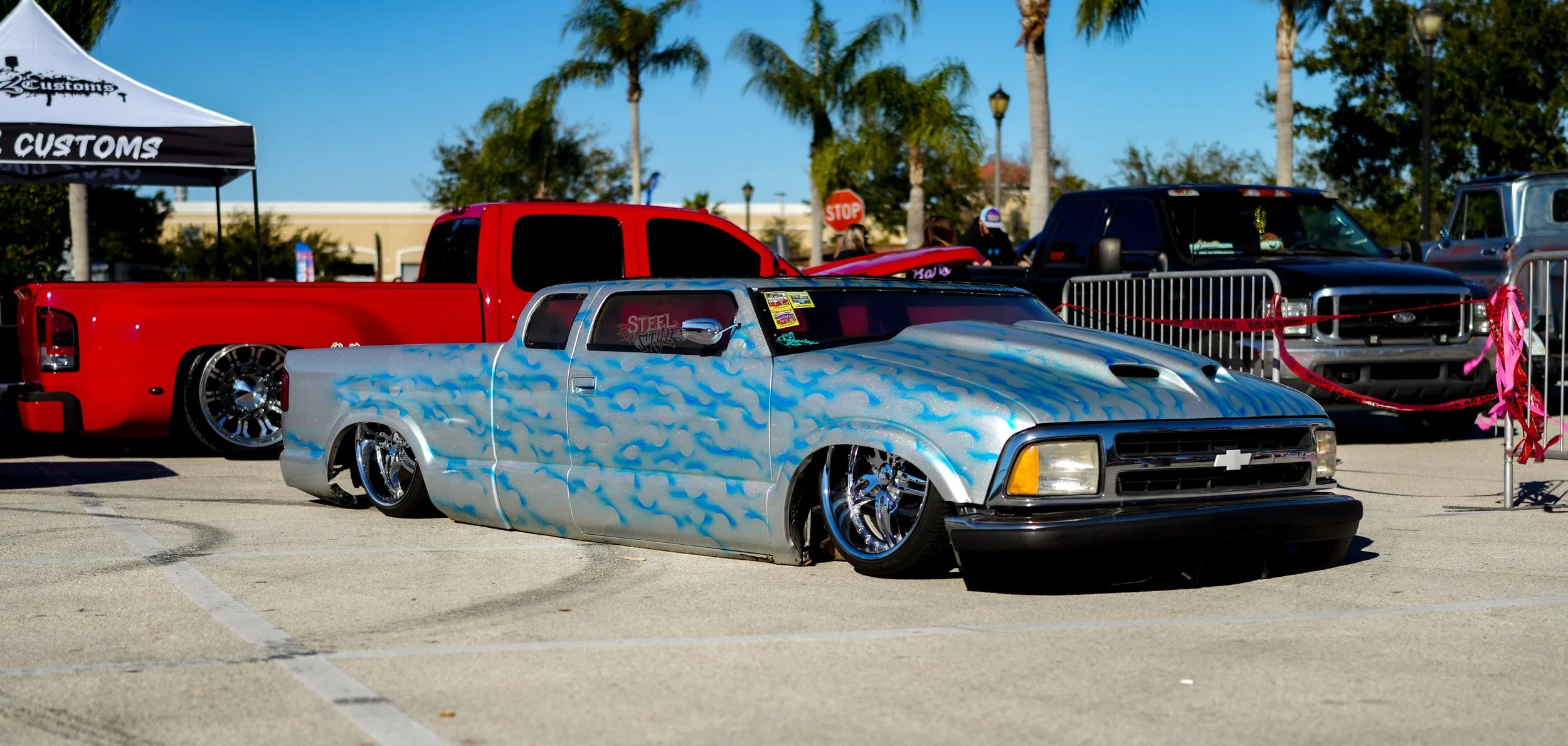 Custom lowrider truck with blue and white flame paint job, chrome rims, and lowered suspension at car show with other vehicles in background under a clear blue sky.