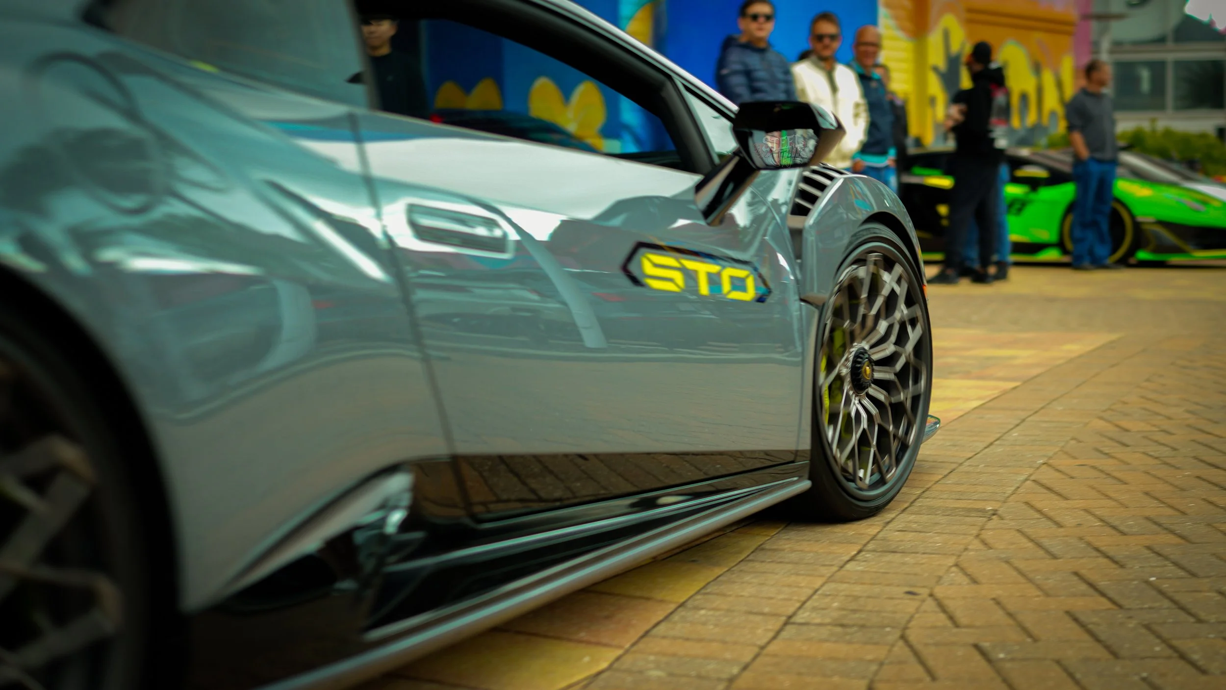 Close-up of a sleek silver sports car with a yellow 'STOP' sticker on the side, parked on a brick pavement at an outdoor car show. In the background, several people and a vivid green supercar are visible.