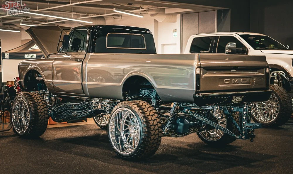 A custom GMC pickup truck with a metallic body, elevated on oversized wheels and suspension, displayed at an indoor auto show.