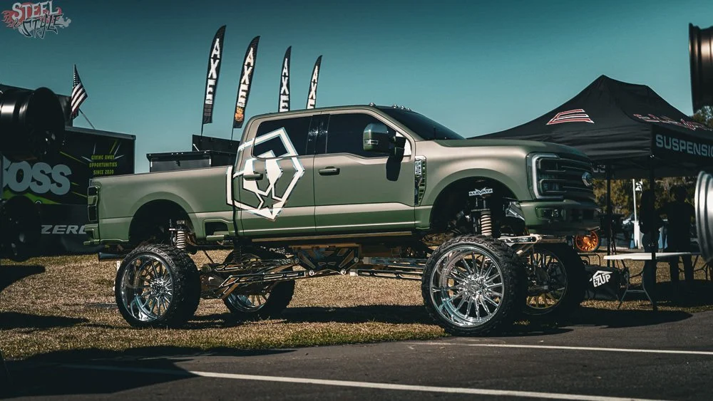 A lifted Ford pickup truck with oversized chrome wheels, parked on display at an outdoor event. The truck has a matte green finish and the logo of a wolf's head on the side. There are banners and a black canopy tent nearby.