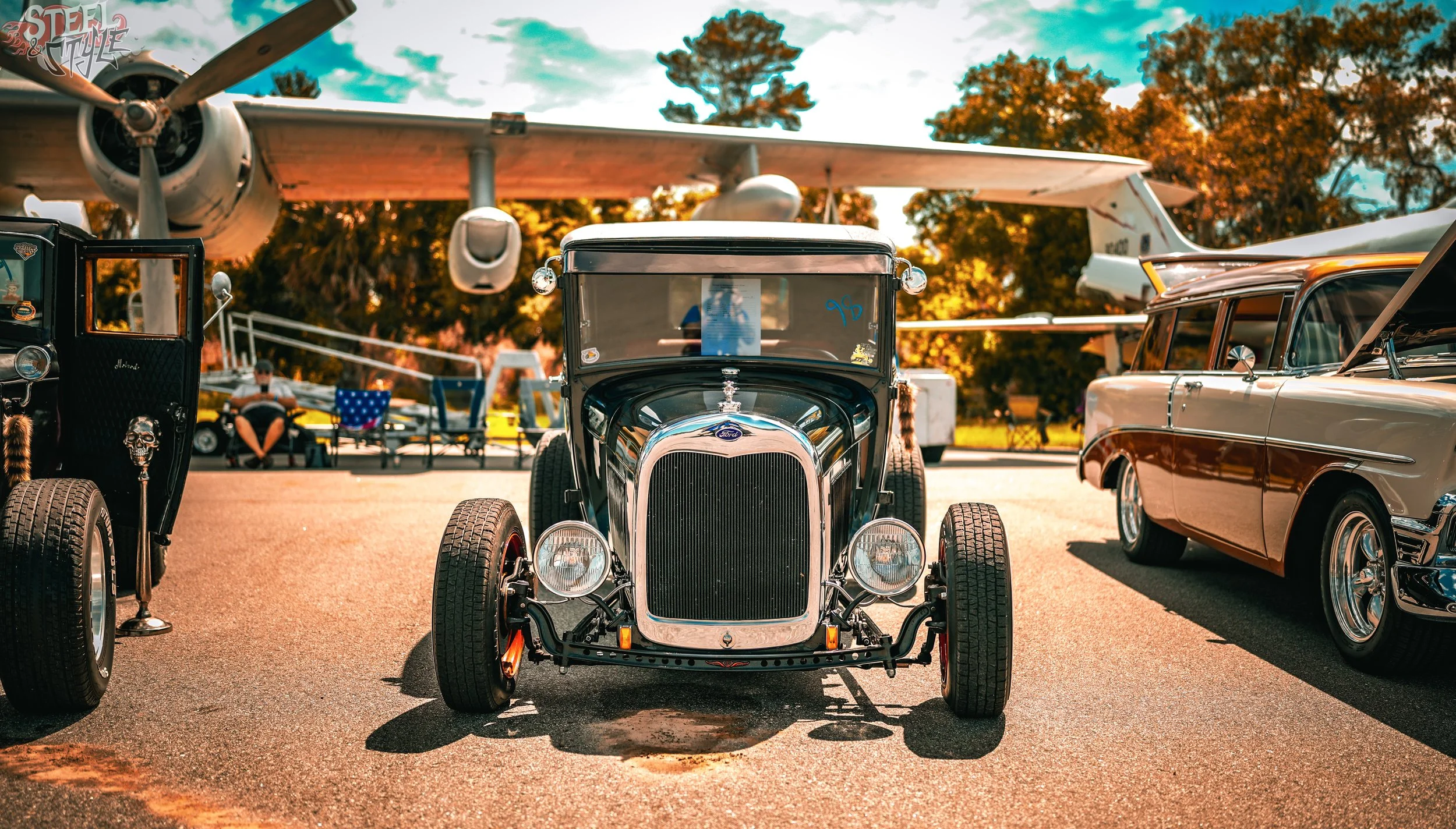 Vintage cars parked outdoors with a large airplane overhead at an outdoor event during autumn.