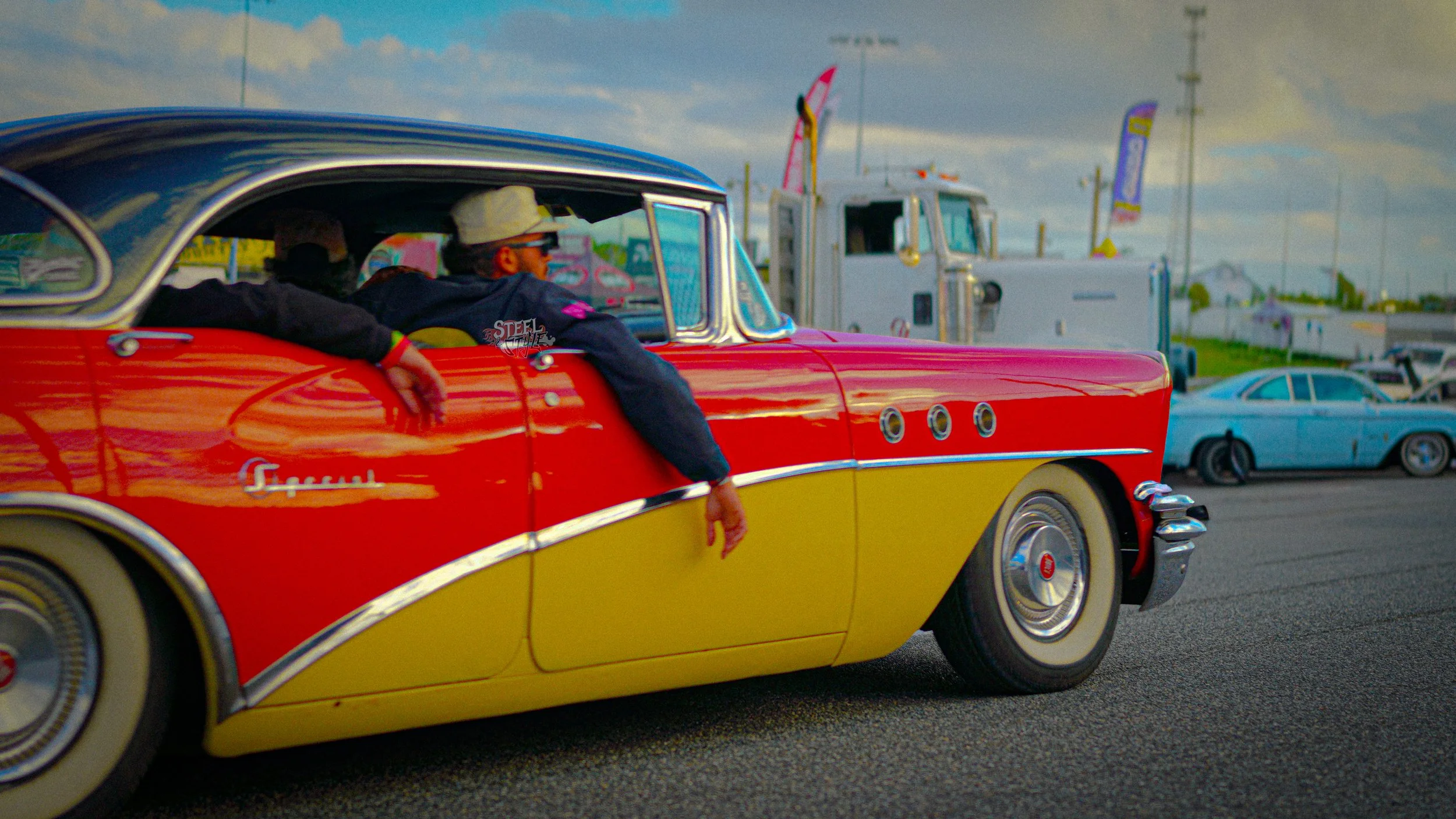 People riding in a vintage red and yellow Cadillac at a car show with other classic cars and trailers in the background.
