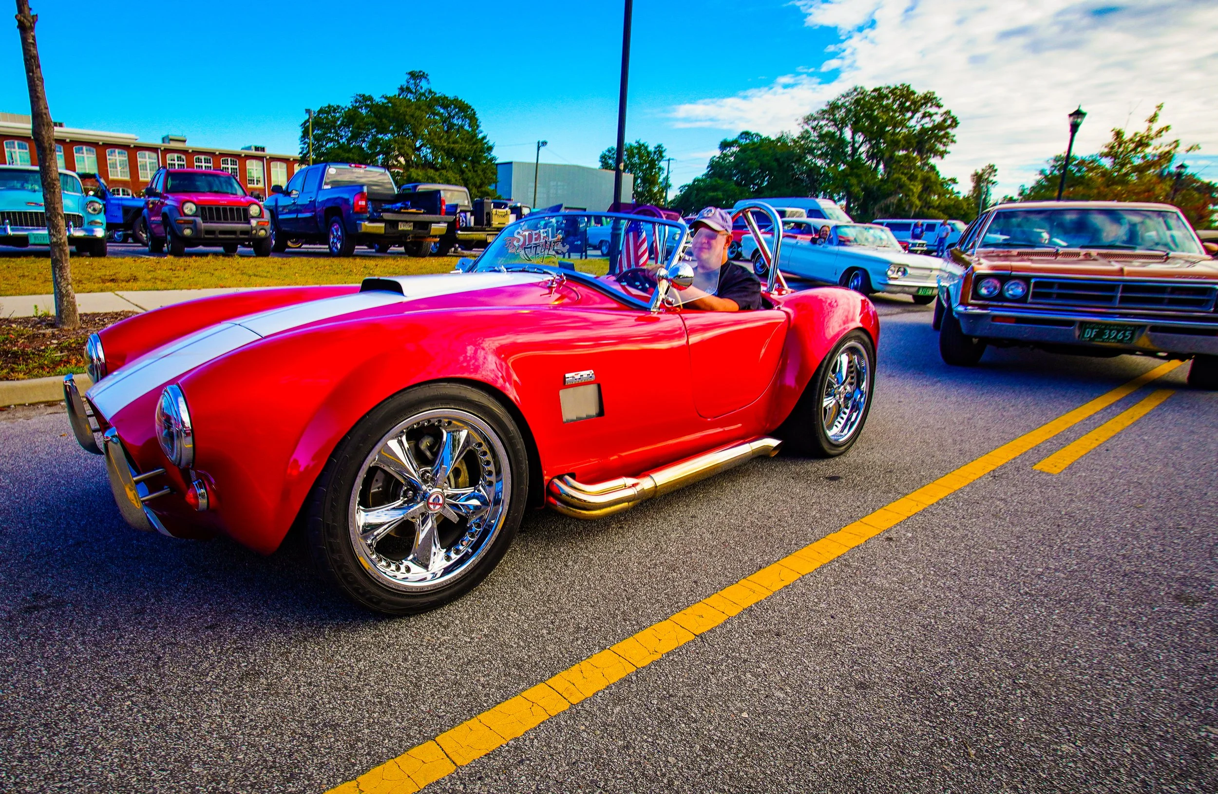A red vintage sports car with white racing stripe, chrome wheels, and exhaust pipes parked on a city street during a classic car show, with other vintage cars and trucks in the background and a man driving.