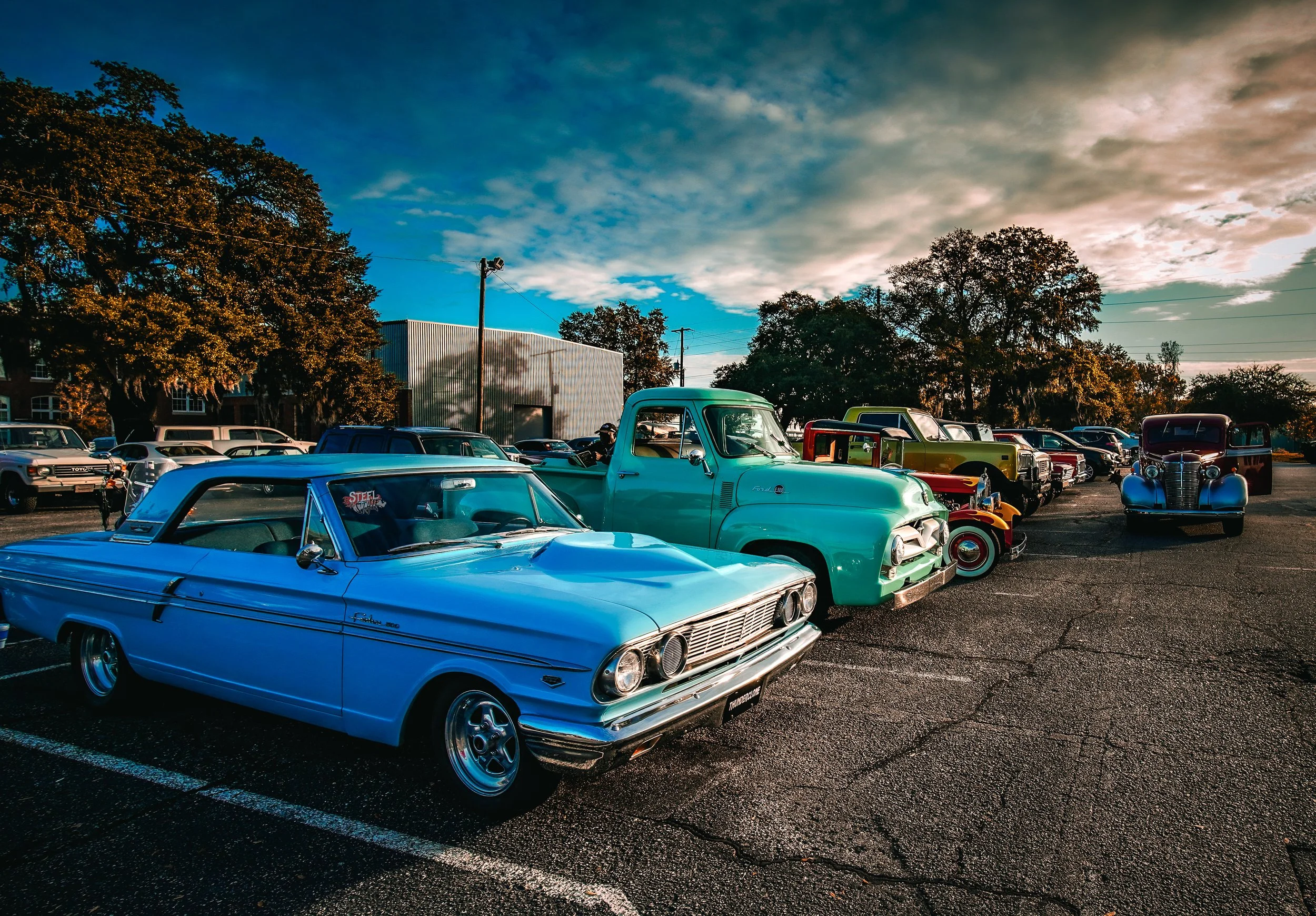 A row of vintage cars parked in a lot with trees and a cloudy sky in the background during sunset.