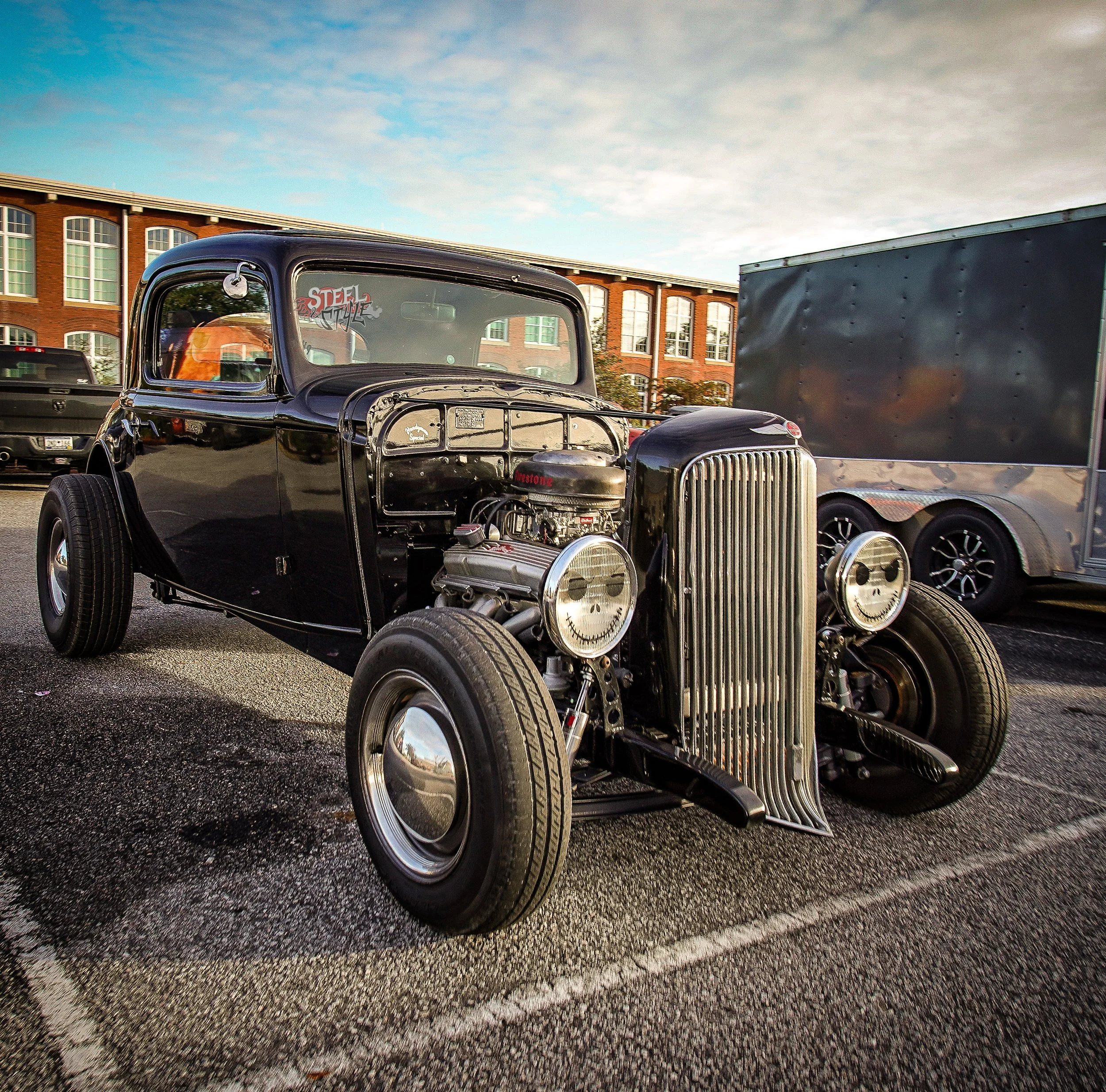 A black vintage car with an exposed engine, parked in a lot with other vehicles and a brick building in the background during the day.