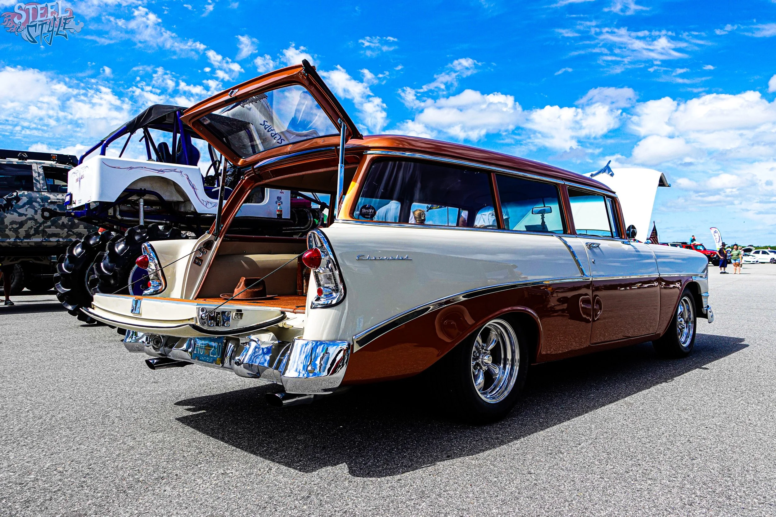 A vintage Chevrolet station wagon with a two-tone paint job in cream and brown, with chrome accents, parked at a car show. The rear hatch is open revealing a racing vehicle mounted inside. The sky is partly cloudy and there are other cars and people 