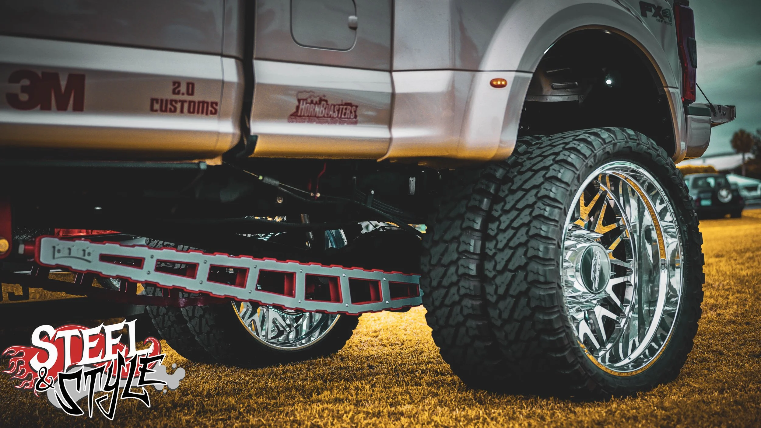 Close-up of a lifted pickup truck with large off-road tires and shiny chrome wheels, parked on grass during sunset, with an off-road racing logo 'Steel & Style' in the bottom left corner.