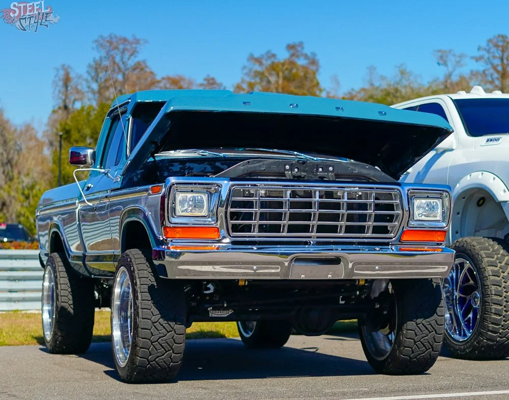 A lifted vintage Ford pickup truck with a blue exterior, large off-road tires, and a chrome grille, parked outdoors with its hood open.