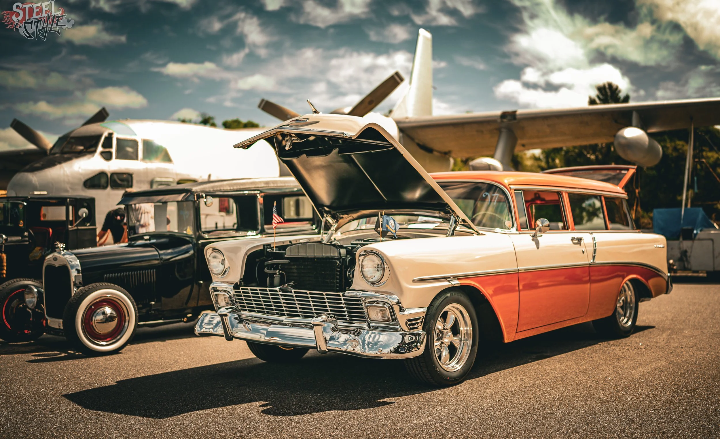 A vintage car with its hood open on display at an outdoor event, with other classic cars and an aircraft in the background under a partly cloudy sky.