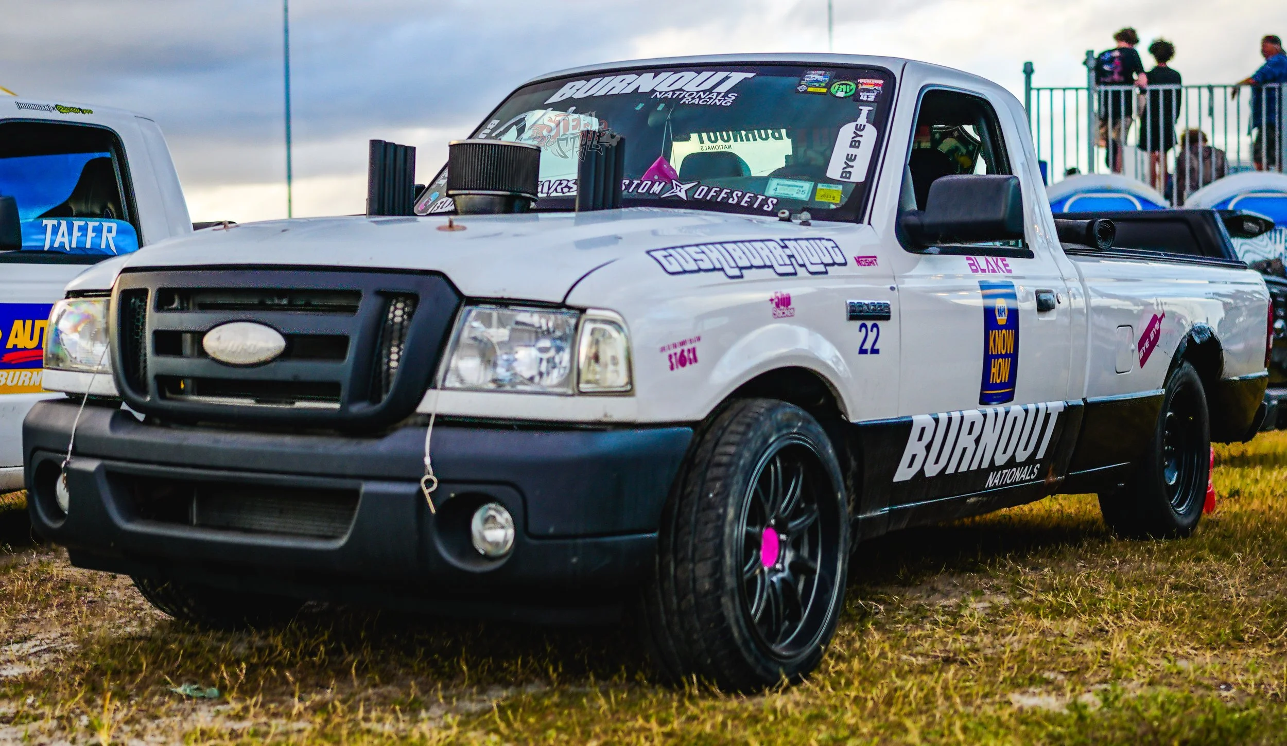 White race truck with various decals and stickers parked on grass, with a black front grille, black wheels, and a black rear bed cover, at a racing event with spectators in the background.