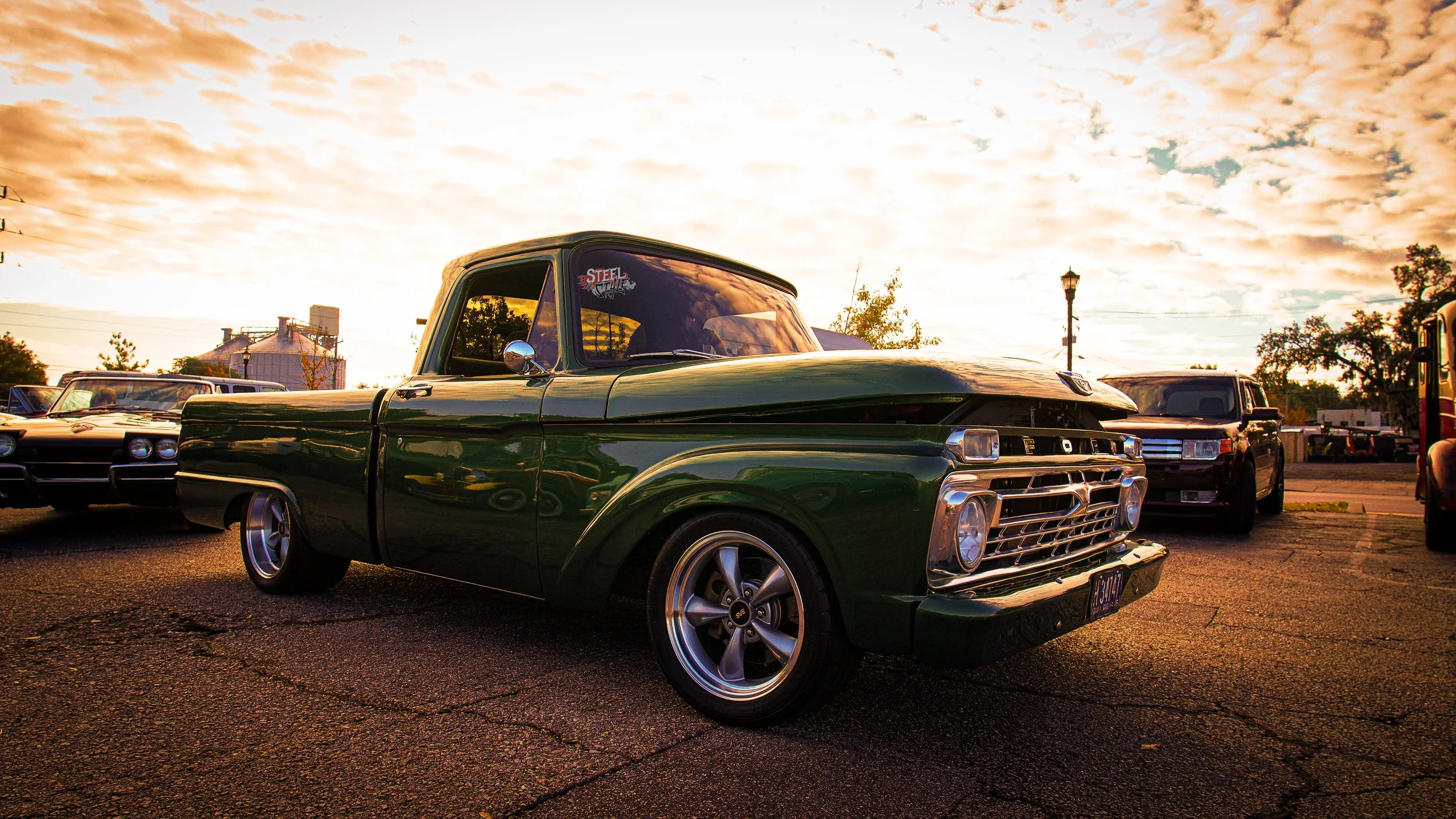 A vintage green pickup truck parked on a cracked asphalt lot during sunset with other cars and a sunset sky in the background.