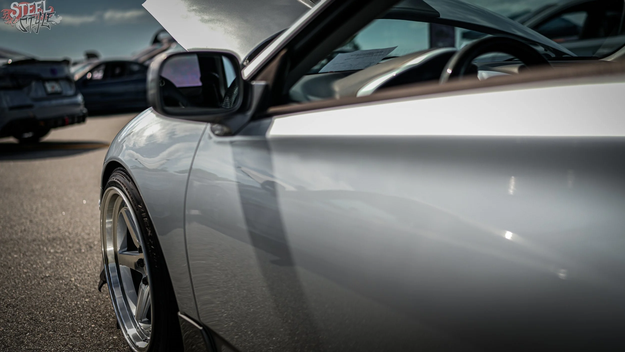Close-up of a silver car with custom rims parked outdoors, with other vehicles blurred in the background.