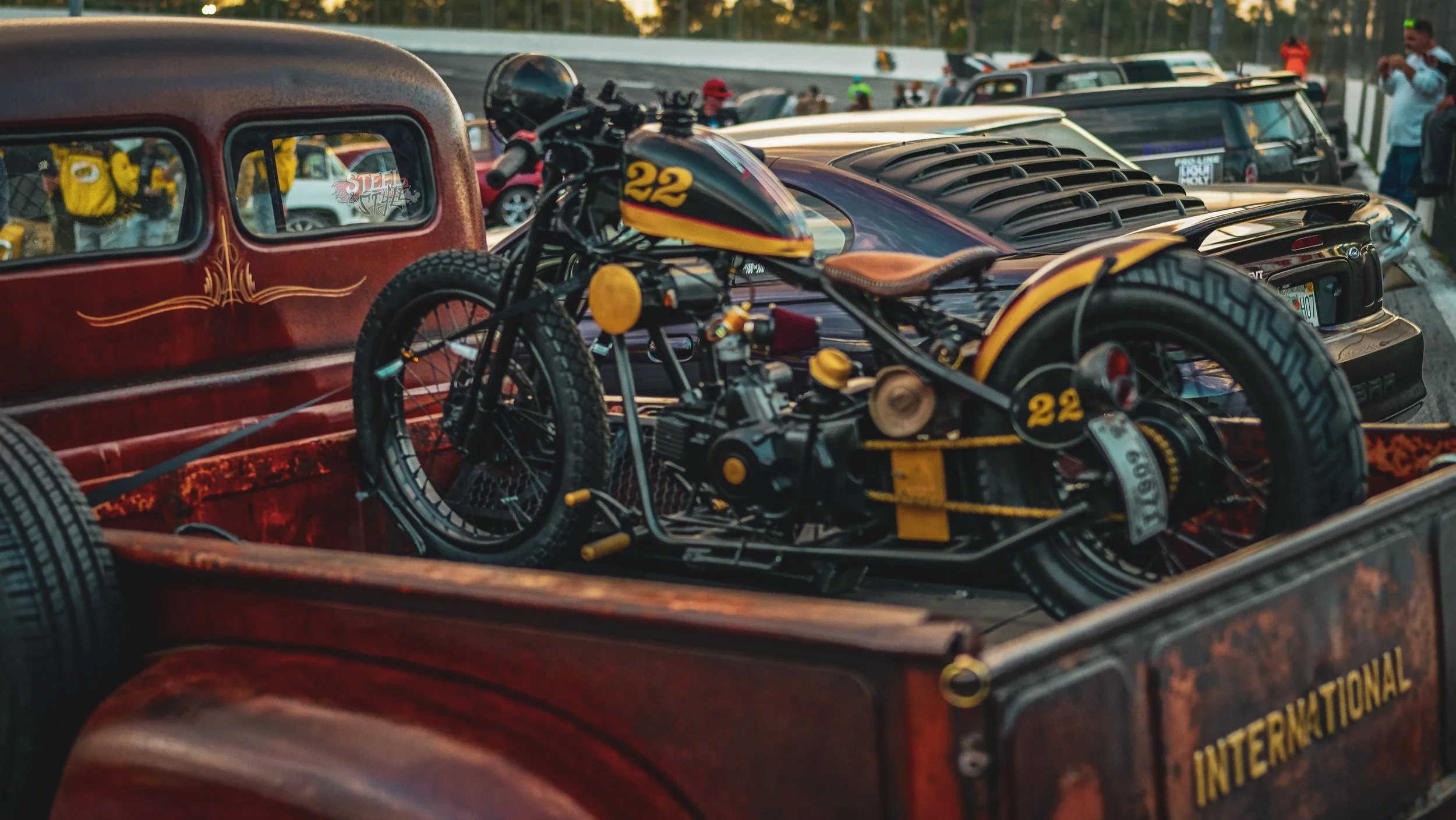 A vintage motorcycle with the number 22 on it, loaded in the back of an old rusty truck at a racing event. Several other cars and people are visible in the background.